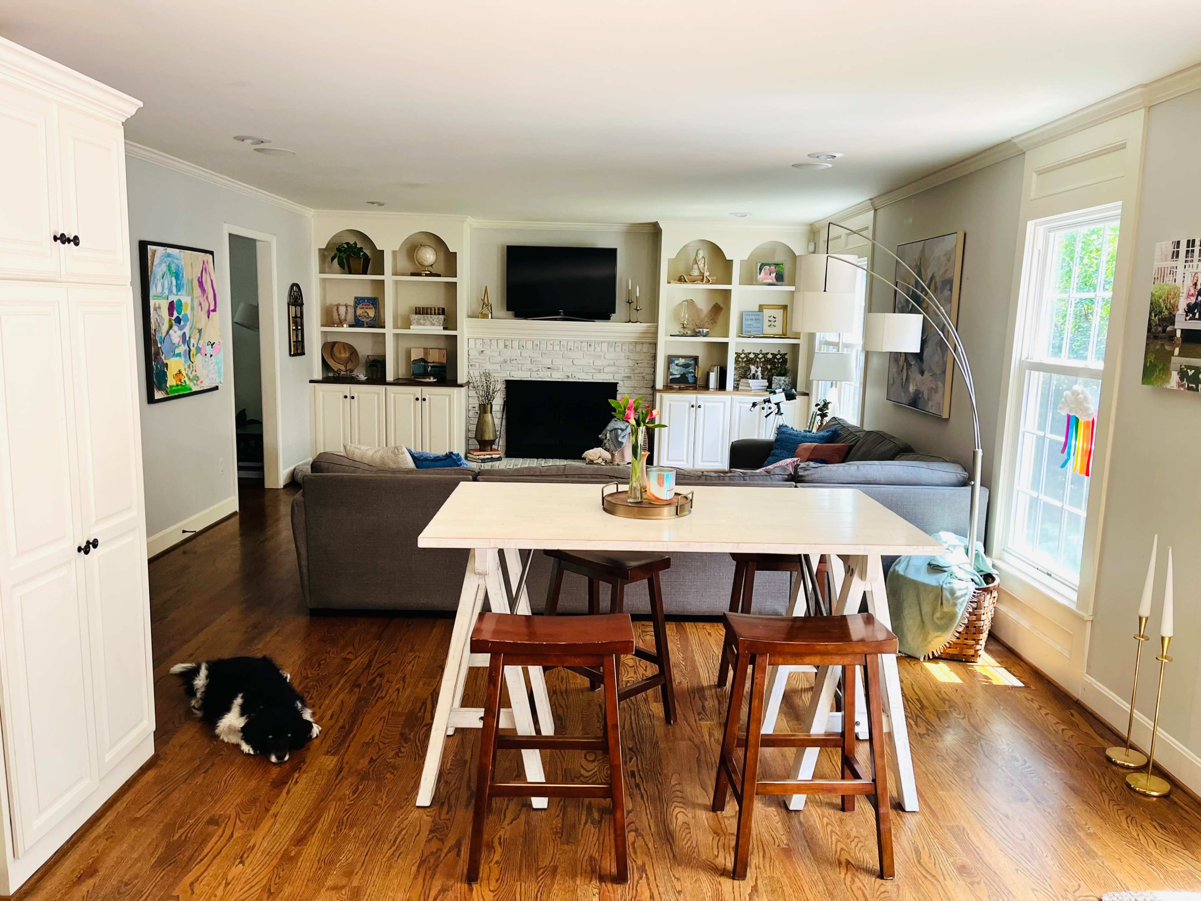 A spacious living room features a gray sofa, a coffee table, and a dining area with two wooden stools, complemented by shelves and a television on the wall.