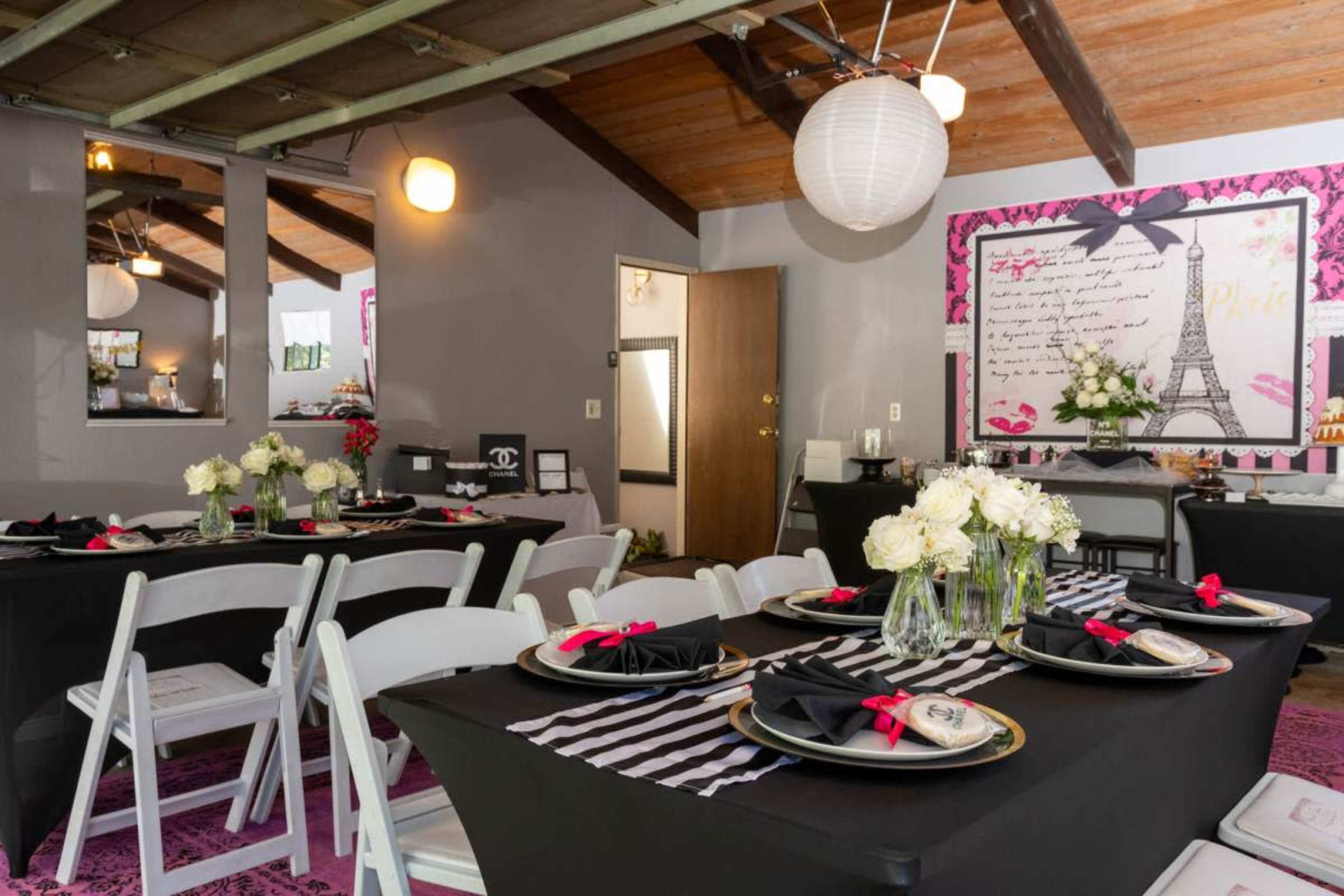 The scene features a beautifully decorated indoor event space with tables set for a gathering, featuring black tablecloths, pink accents, and floral centerpieces.