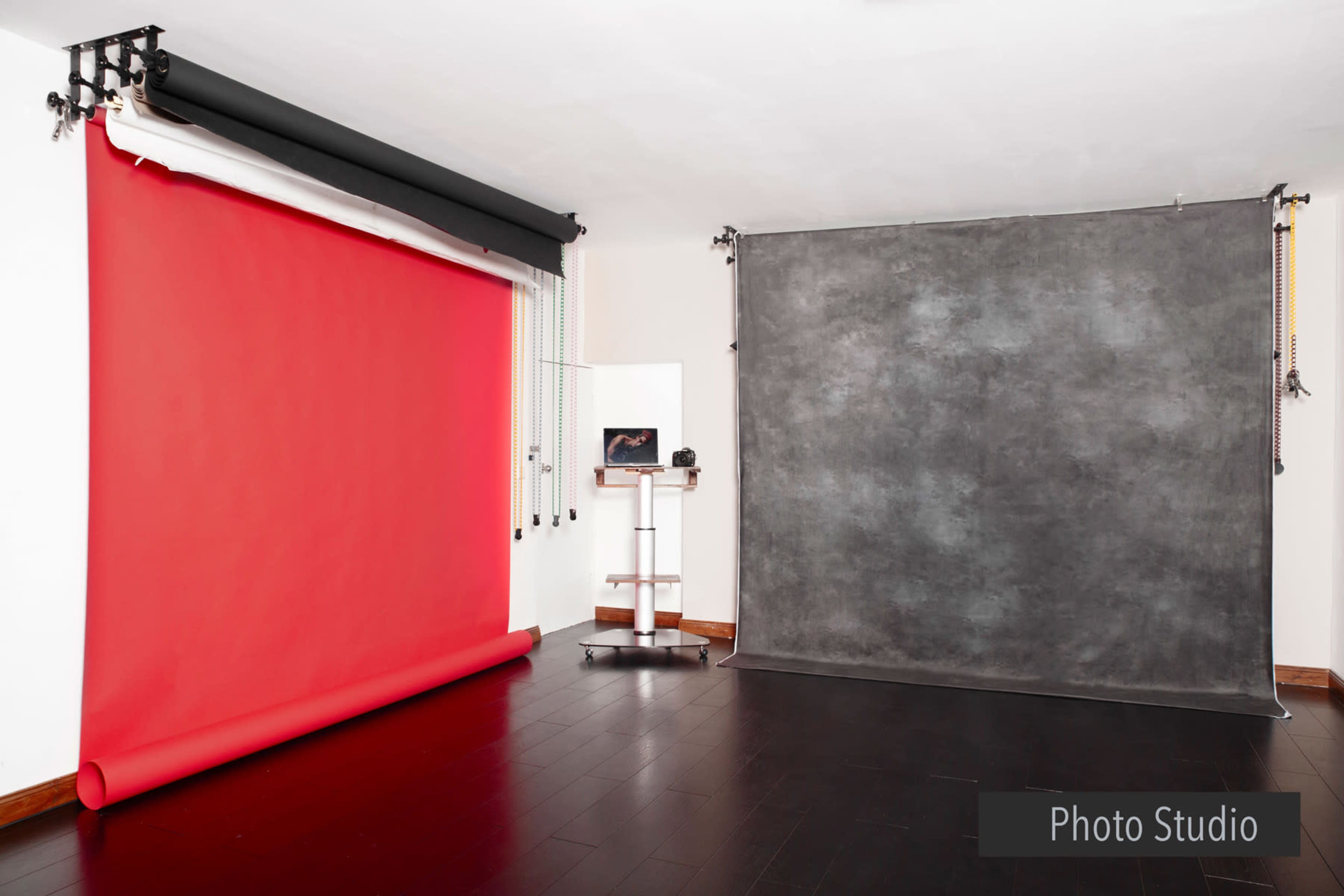 A photo studio with a red backdrop on the left and a gray backdrop on the right, along with a small cart and a computer monitor.