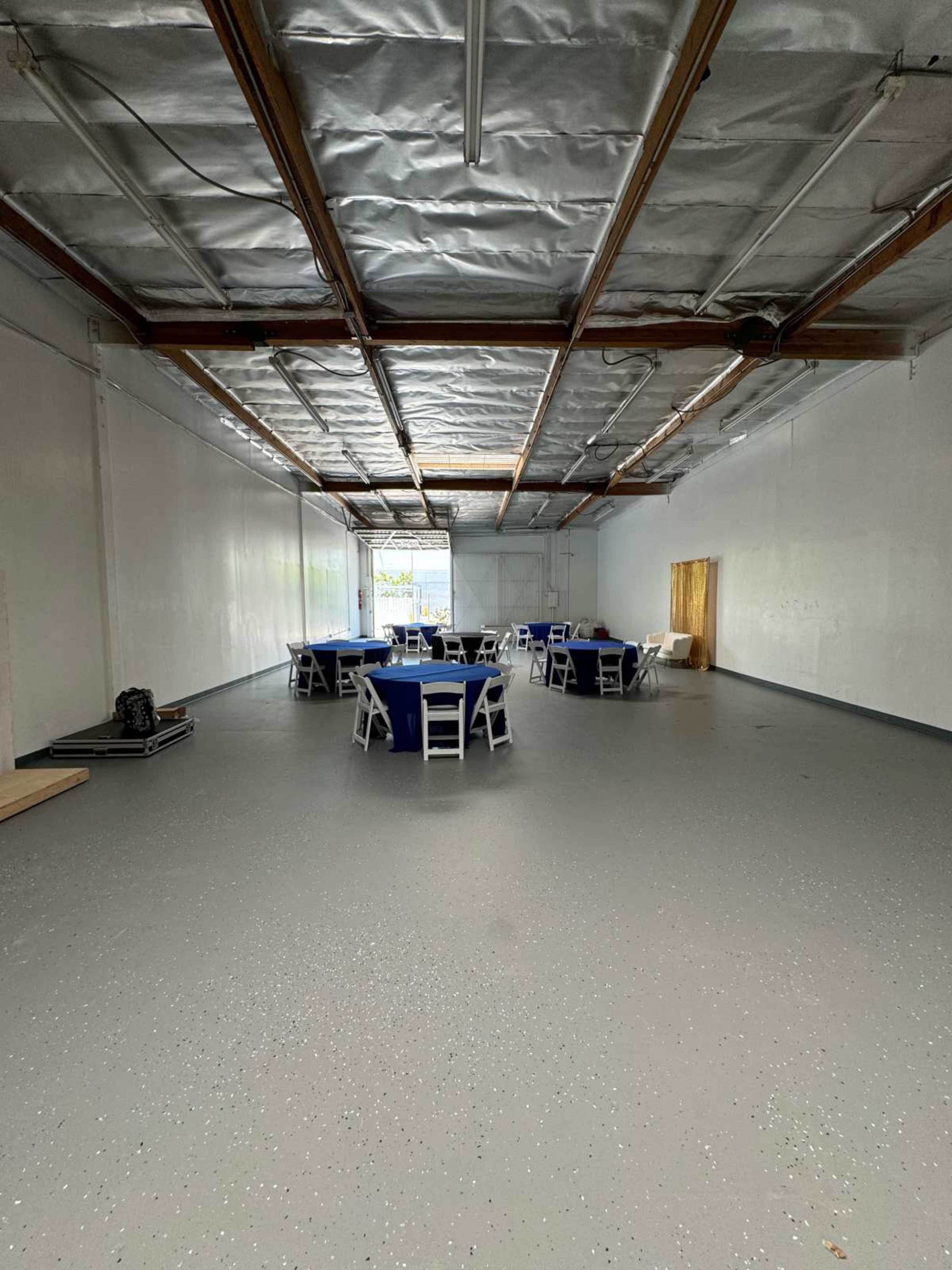 A spacious, empty room with a polished gray floor and several round tables covered with blue tablecloths, surrounded by white chairs.