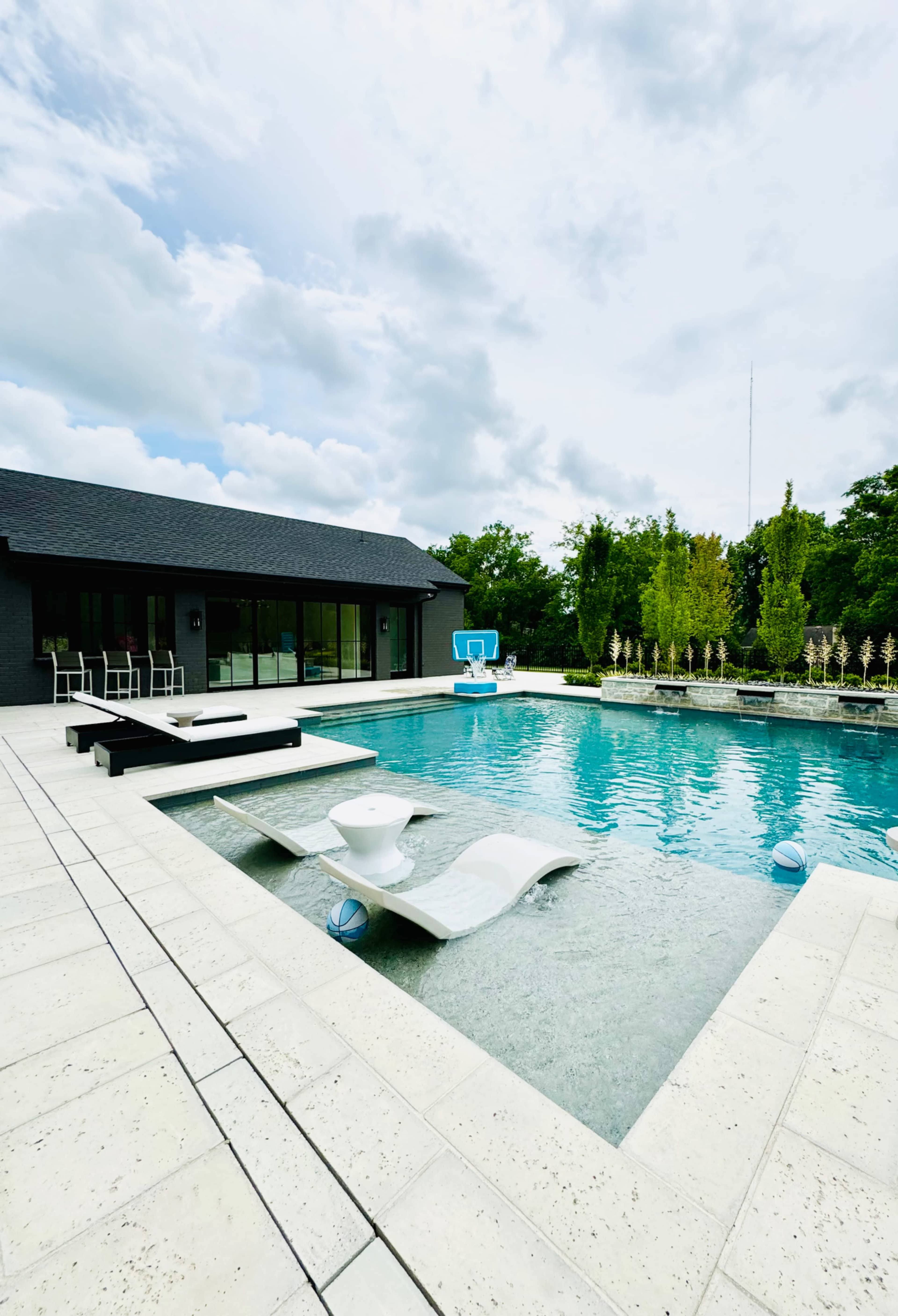 A modern swimming pool surrounded by a patio and a contemporary house in a landscaped outdoor area under a cloudy sky.