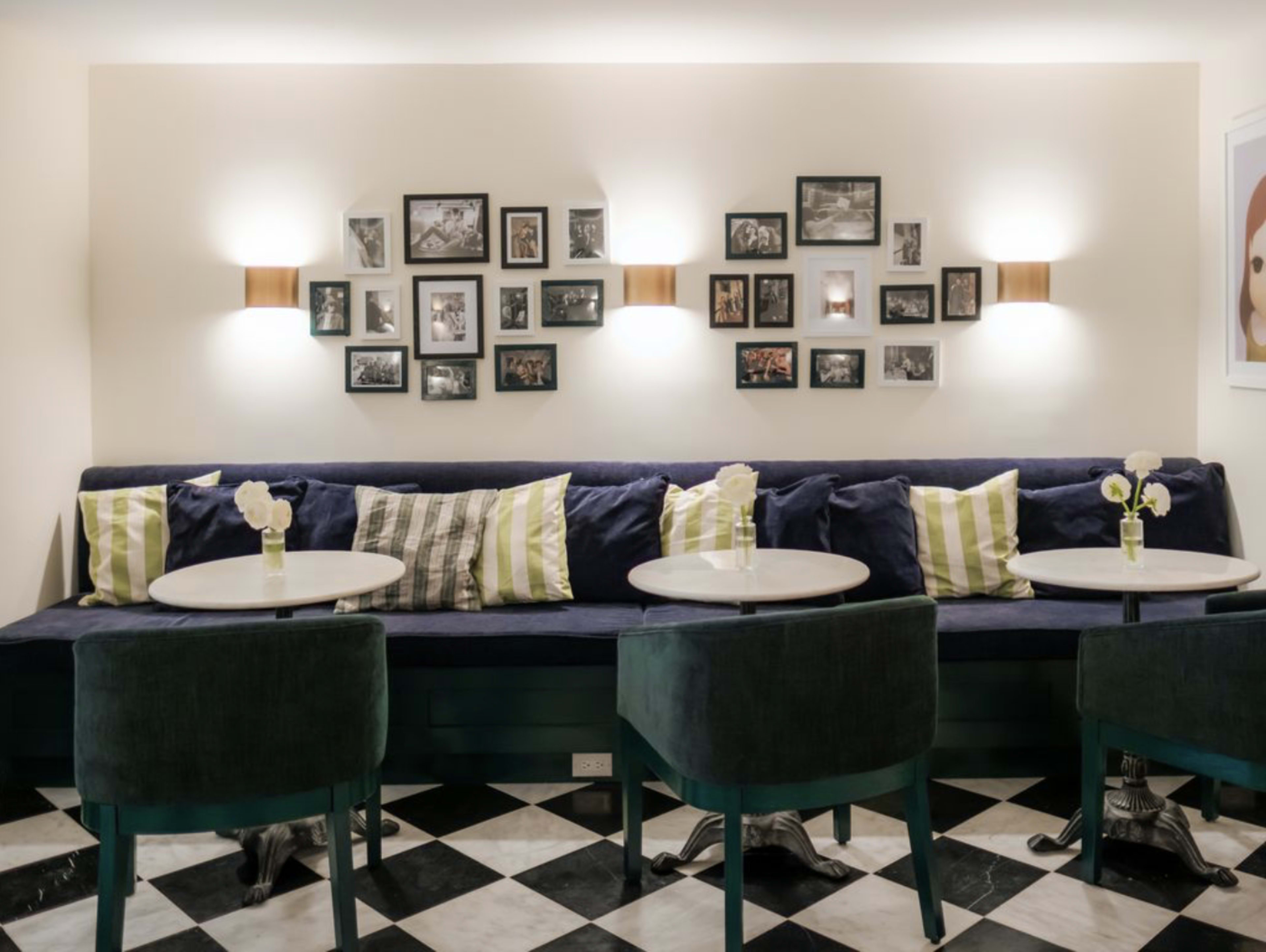 The image shows a cozy seating area with a long blue couch, several small round tables, and framed photographs hanging on the wall above.