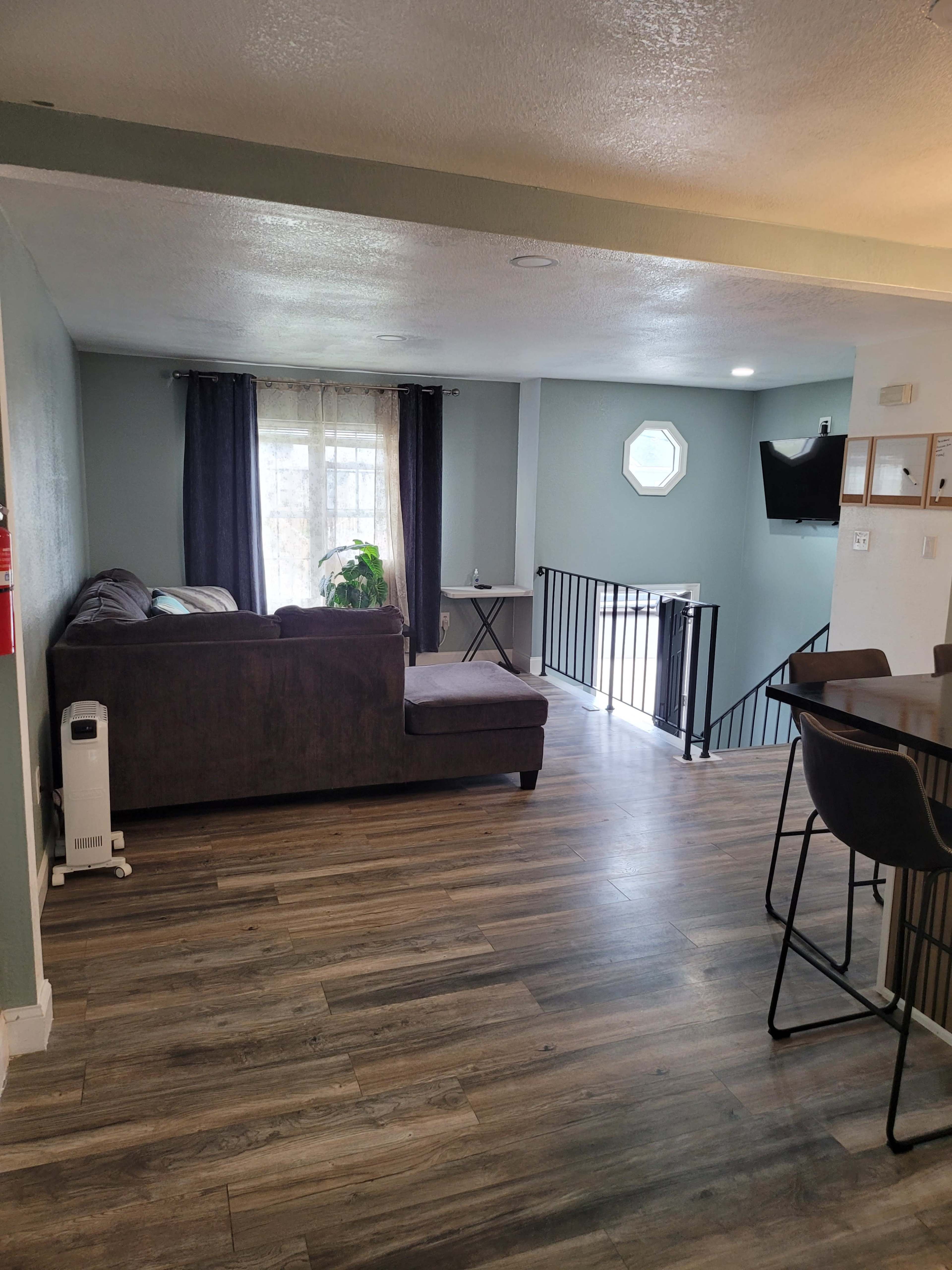 The image shows a living space with a gray sectional sofa, a small table, and stairs leading to another level, all featuring wood-like flooring and light blue walls.