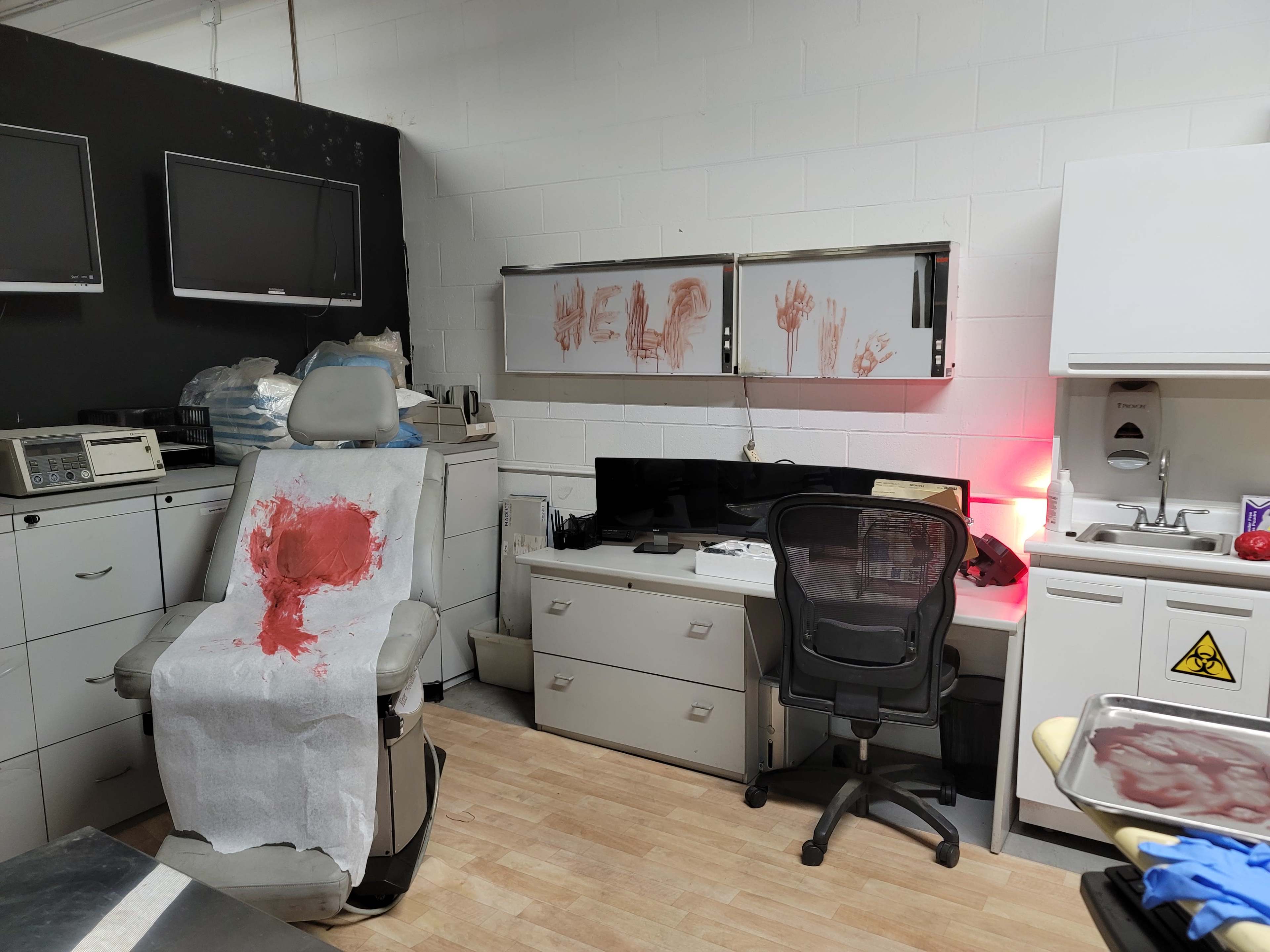 A dimly lit room with medical equipment, a chair stained with red, and handprints on the walls.