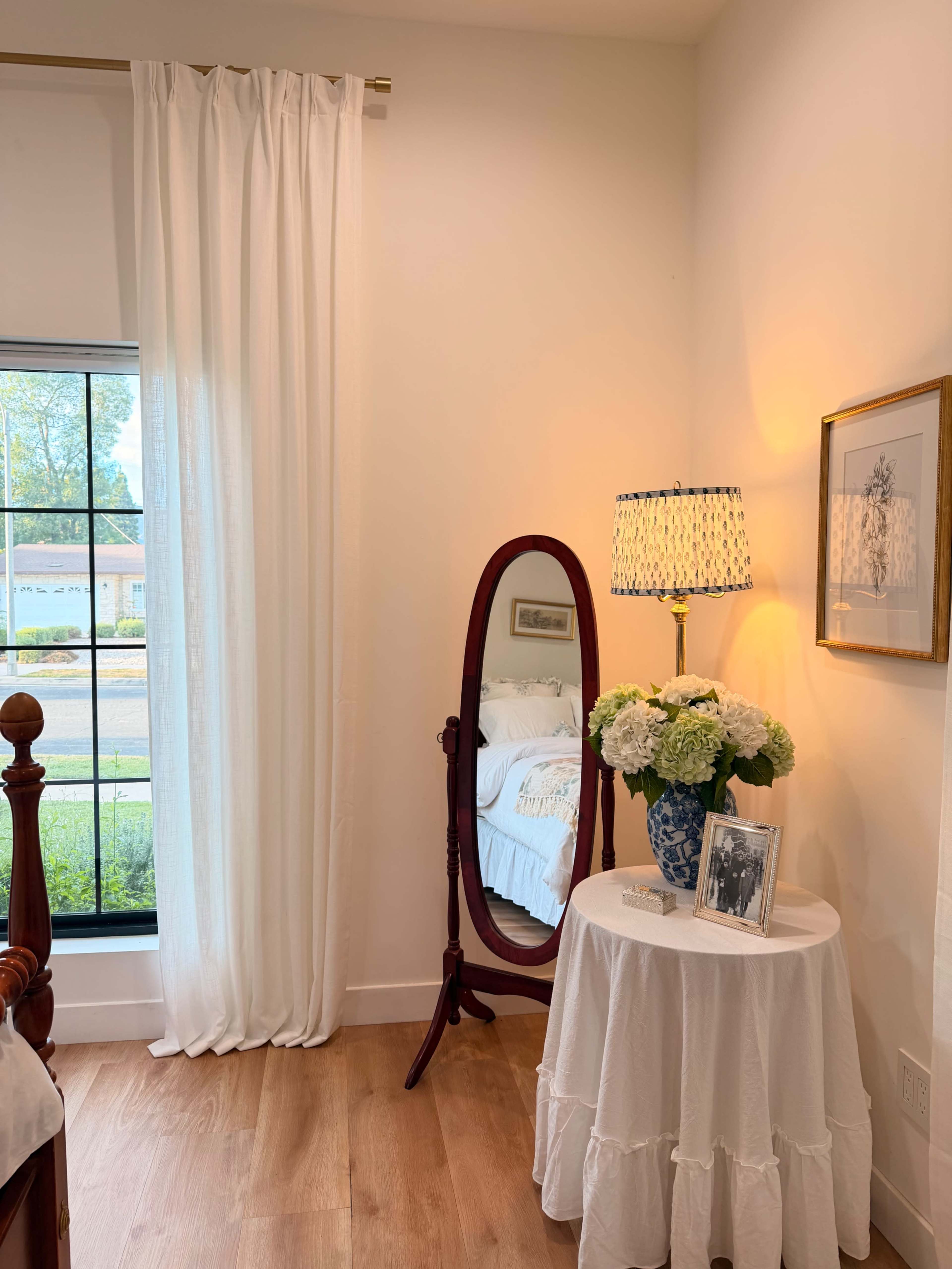 The image shows a corner of a bedroom, featuring a vintage-style mirror on a small round table, accompanied by a lamp and a vase of white flowers.