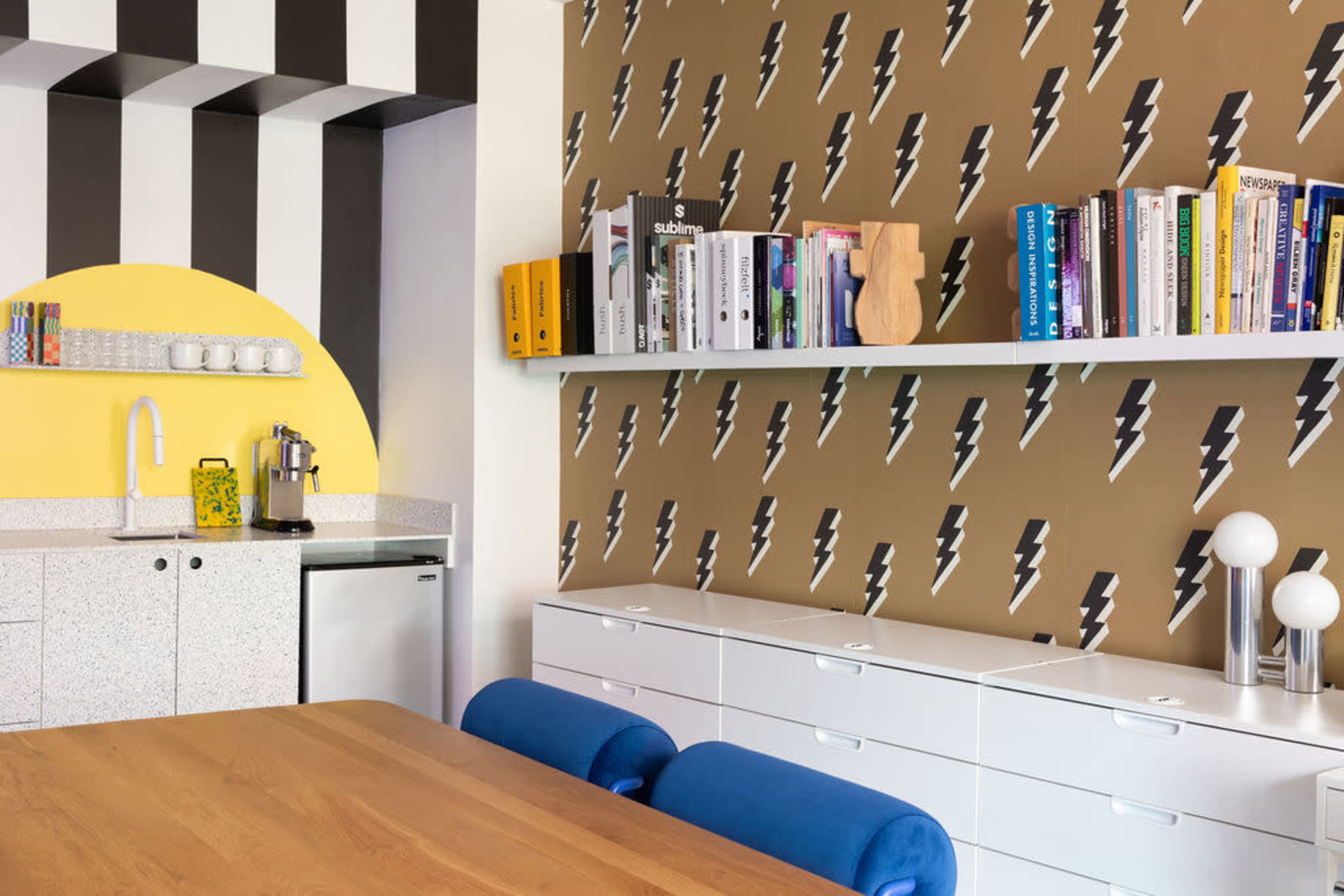 The image shows a modern kitchen with a patterned wall featuring lightning bolts, a wooden dining table, and a white shelf filled with books.