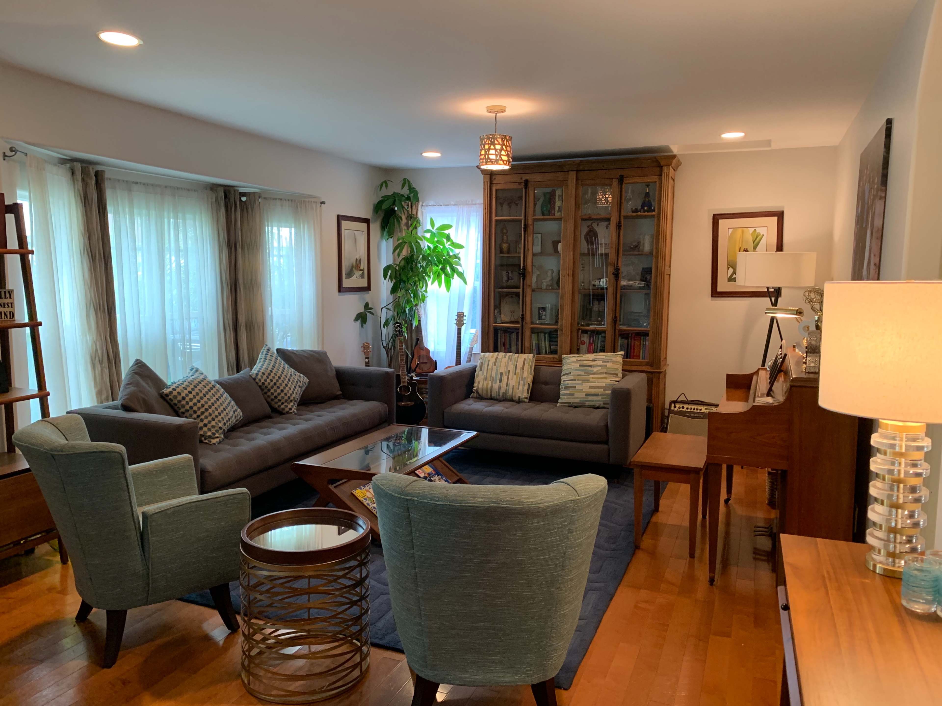 A modern living room features two gray sofas, a glass coffee table, a wooden bookshelf, and a piano, with natural light streaming through the windows.