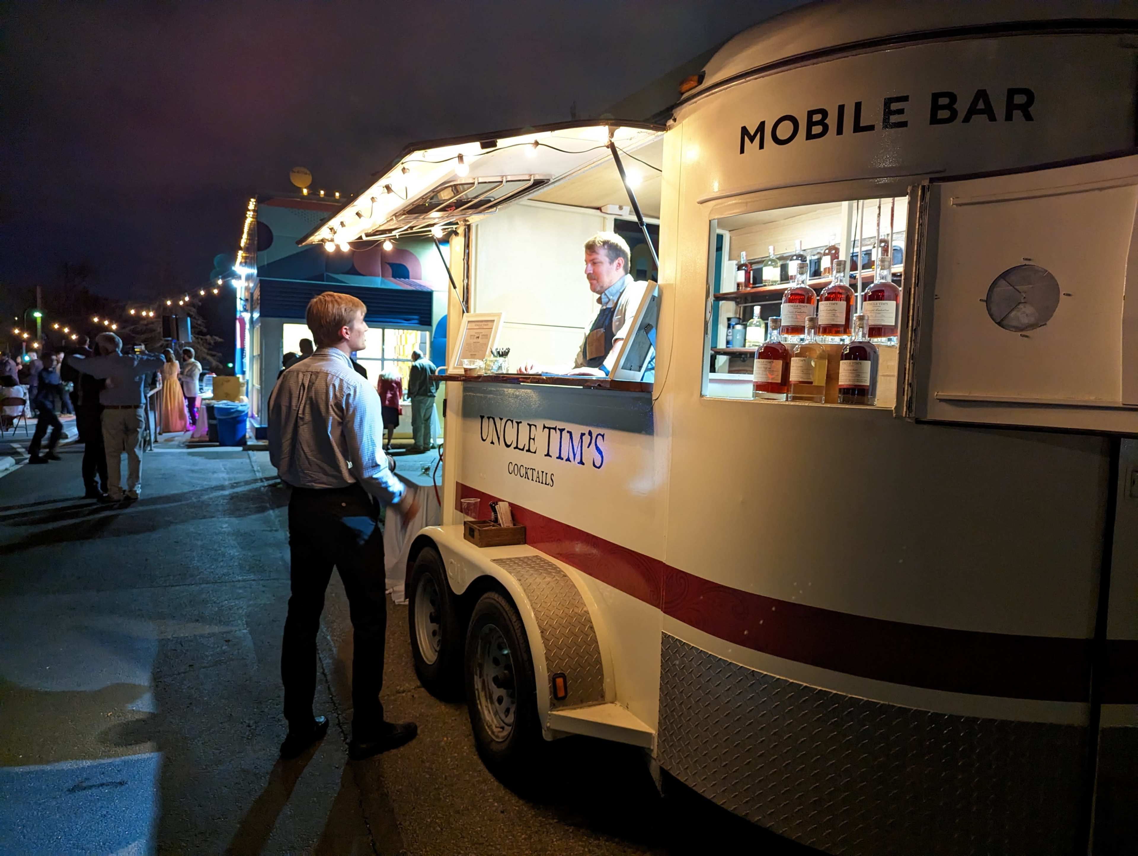 A man in a dress shirt talks to a bartender at a mobile bar during an evening event.