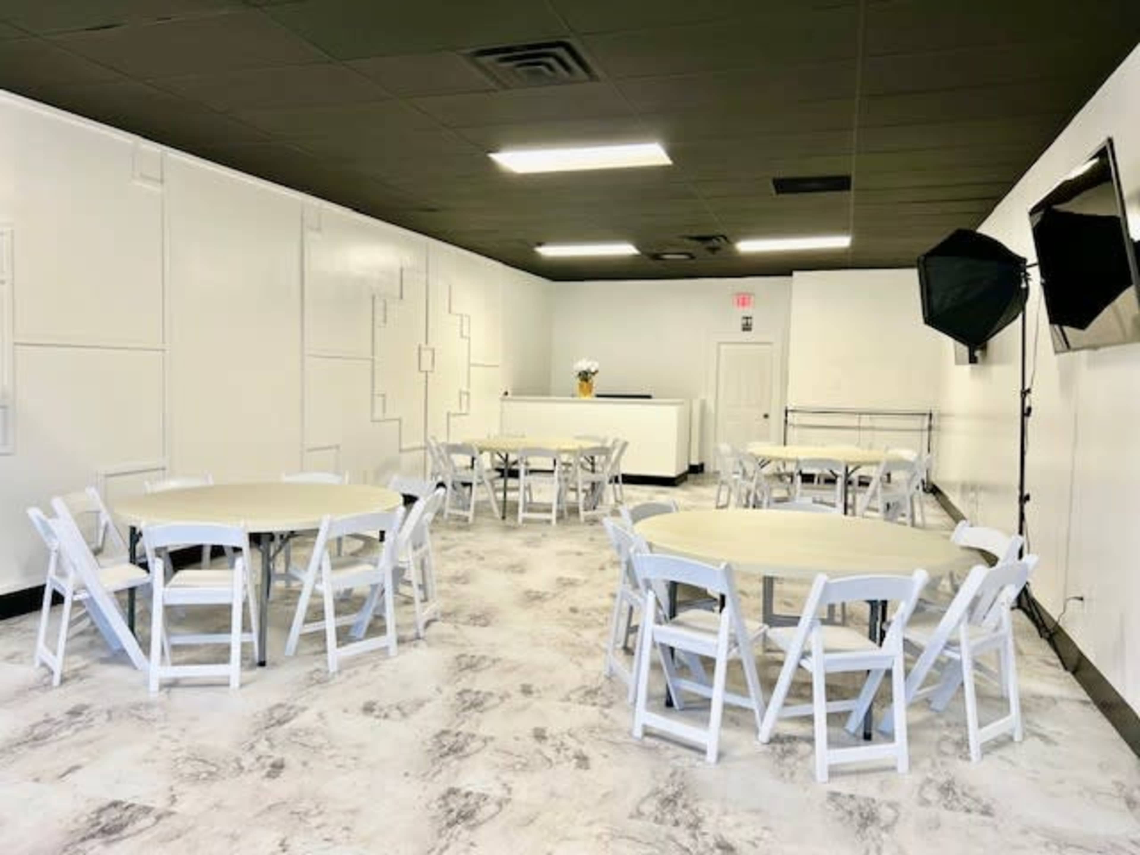 A simple, empty event space with white walls, several round tables with white chairs, and a small stage area at the front.