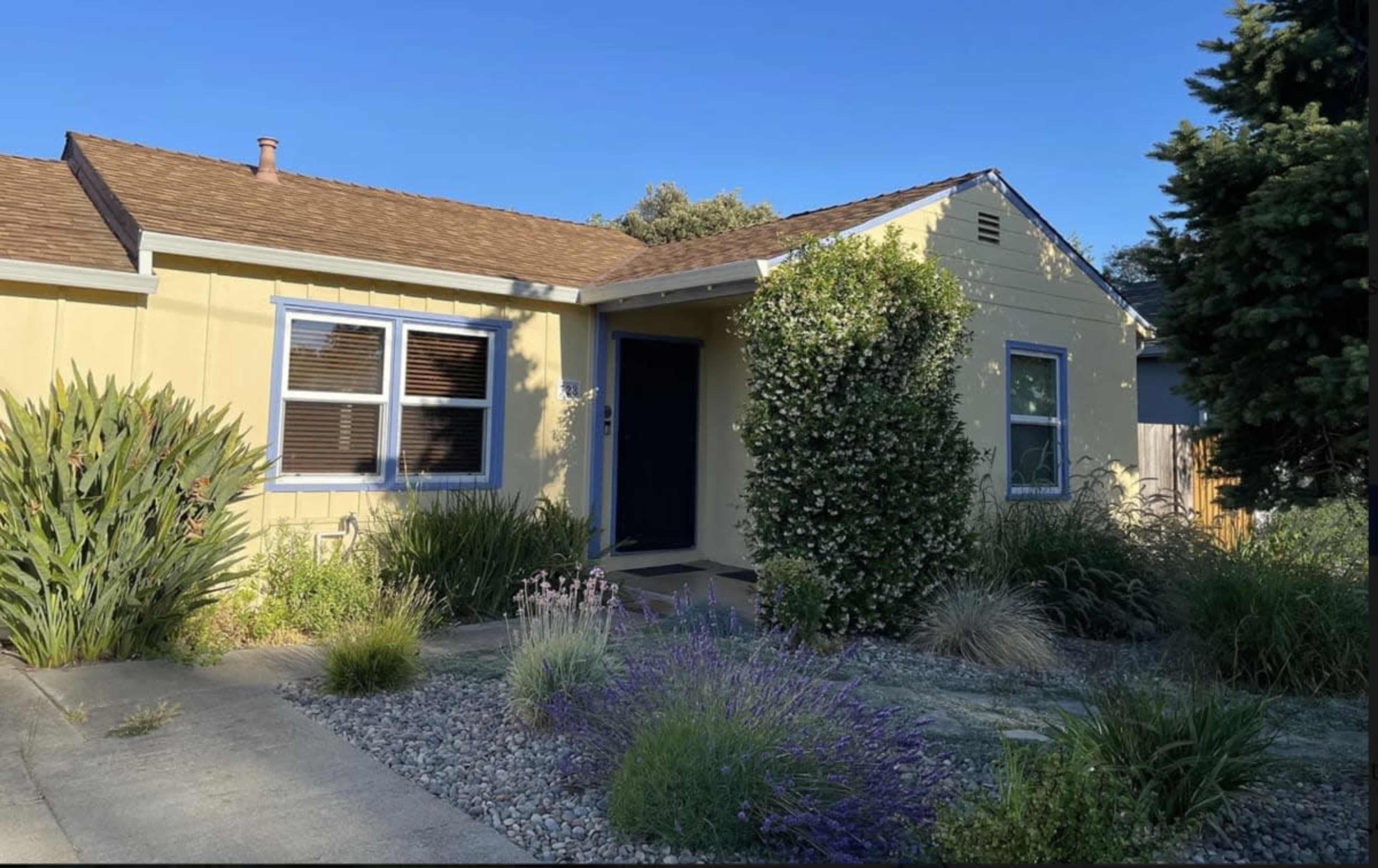 The image shows a single-story yellow house with a brown roof, surrounded by landscaped greenery and a pathway leading to the entrance.
