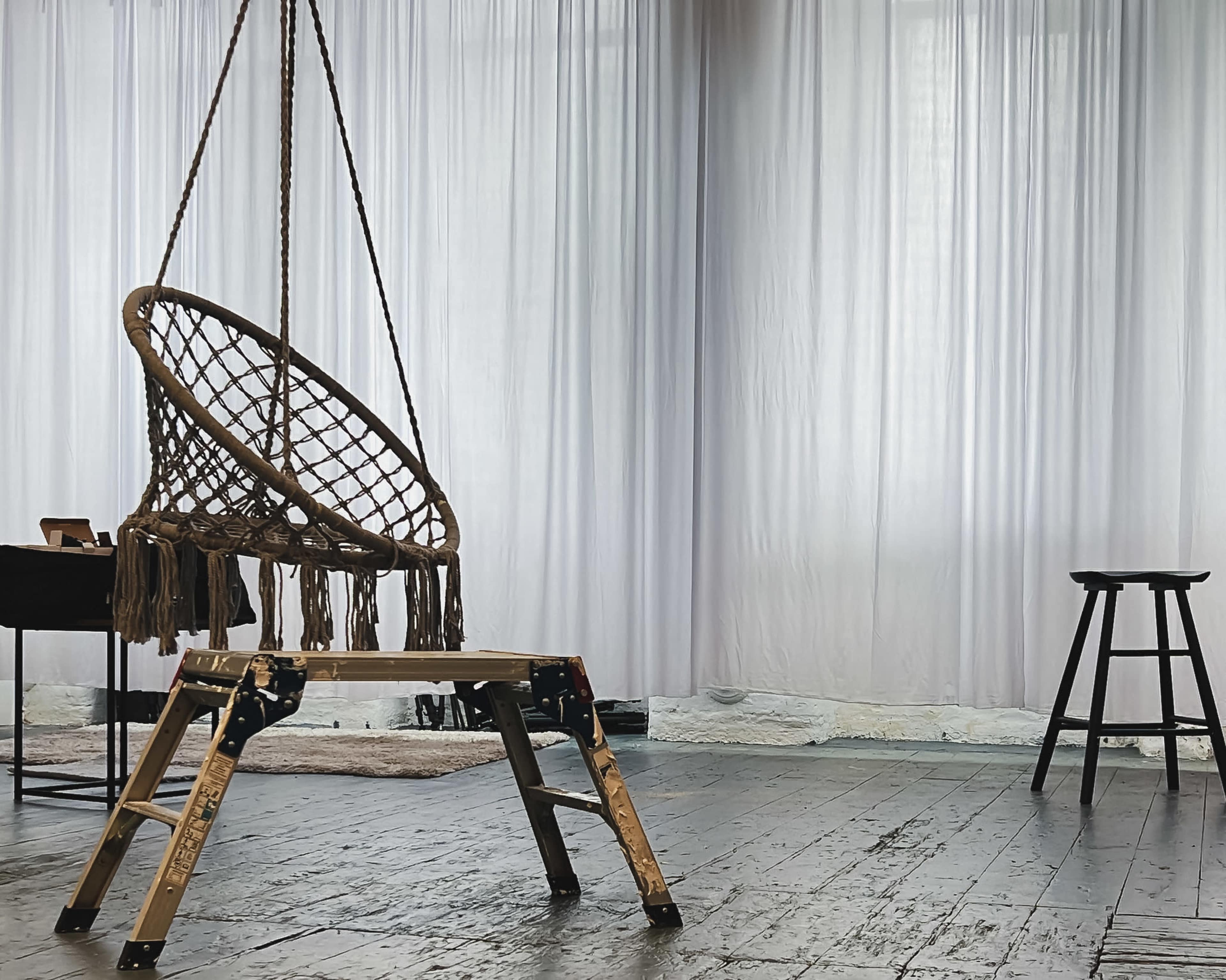 A macramé hanging chair is suspended from a wooden ladder in a spacious, softly lit room with sheer curtains.