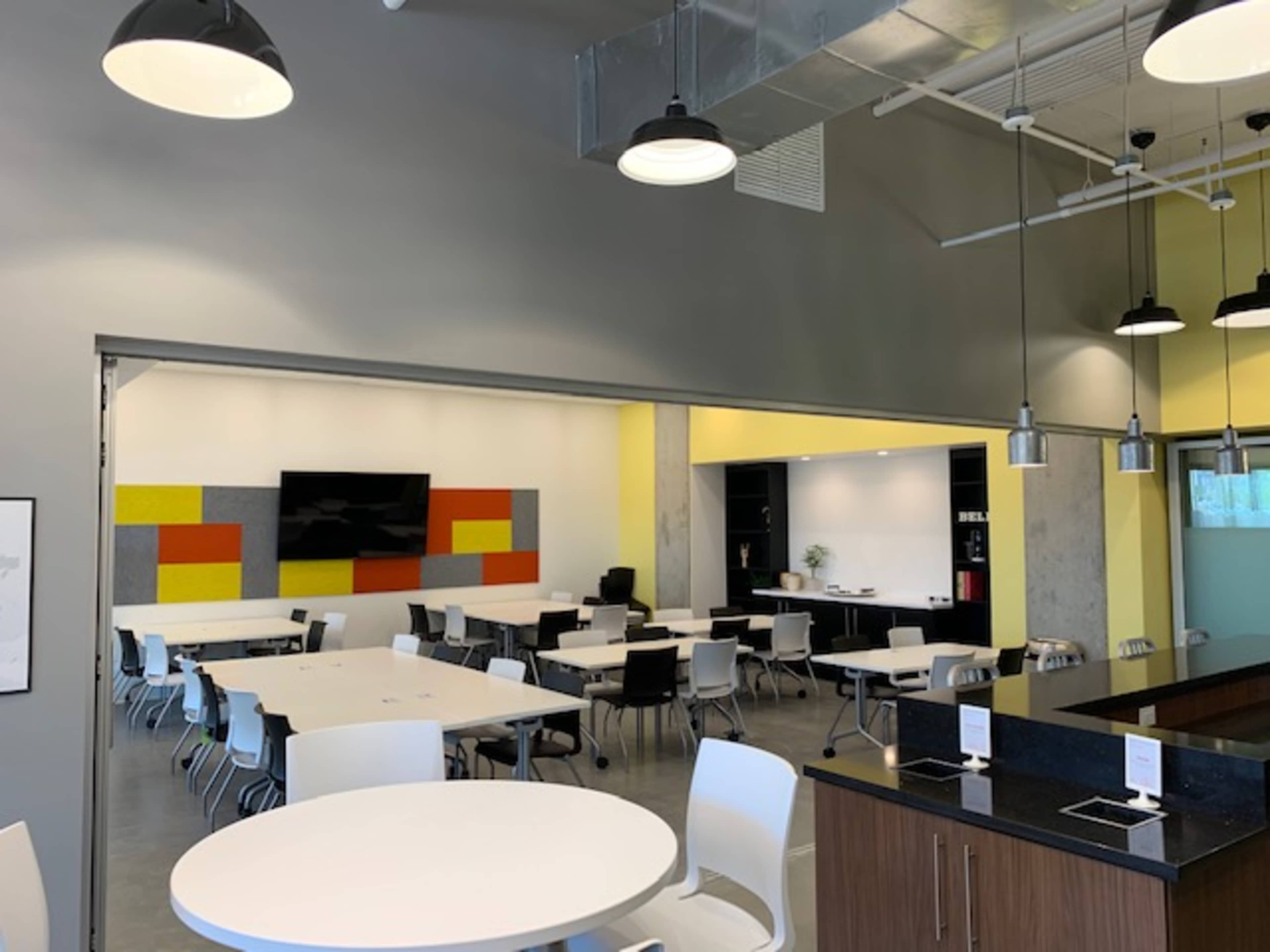 A modern, open-plan dining area with white tables and chairs, a large television screen on the wall, and a neighboring workspace featuring shelves and plants.