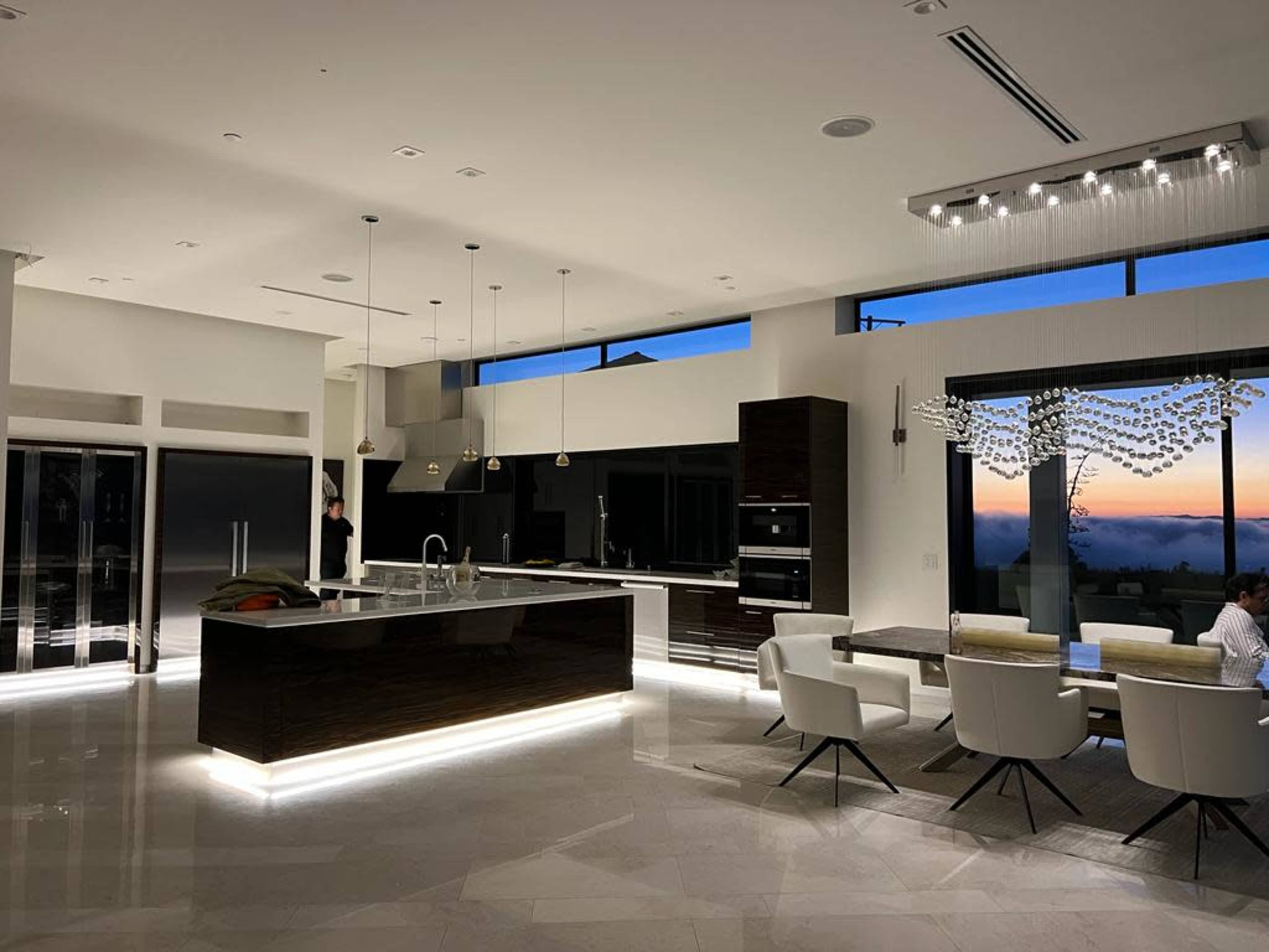 A modern kitchen with a central island, dining area, and large windows displaying a sunset.