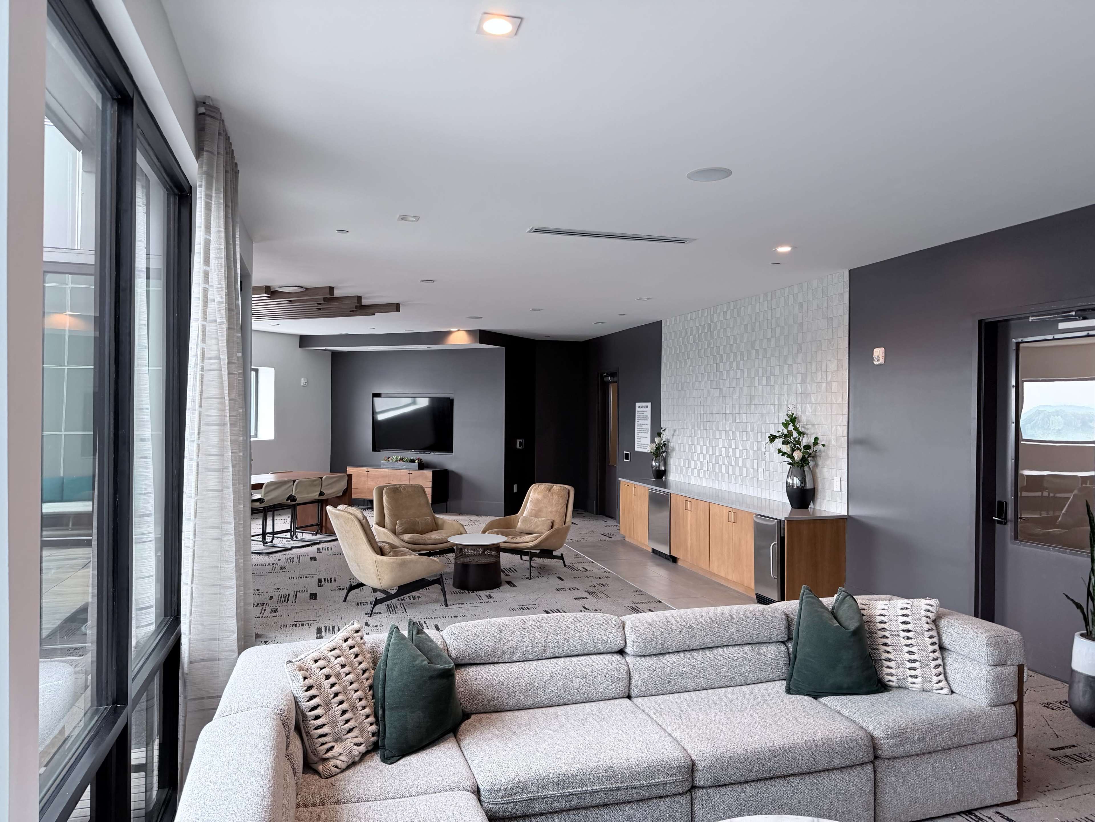 The image shows a modern lounge area featuring a sectional sofa, a coffee table, and lounge chairs arranged around a large television on the wall, with large windows allowing natural light to enter.