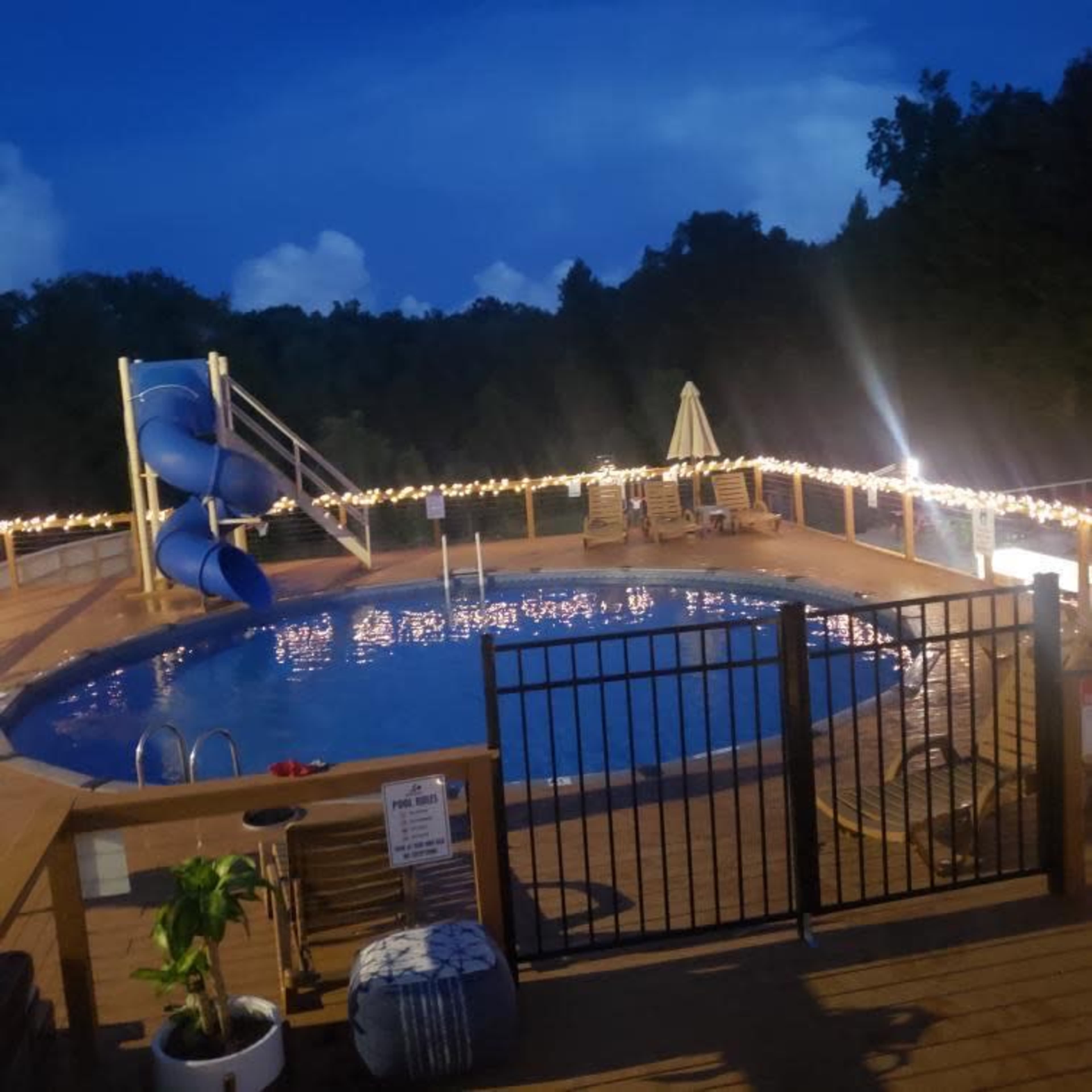 The image shows a pool area at dusk, featuring a blue slide, string lights along the railing, and several lounge chairs around the water.