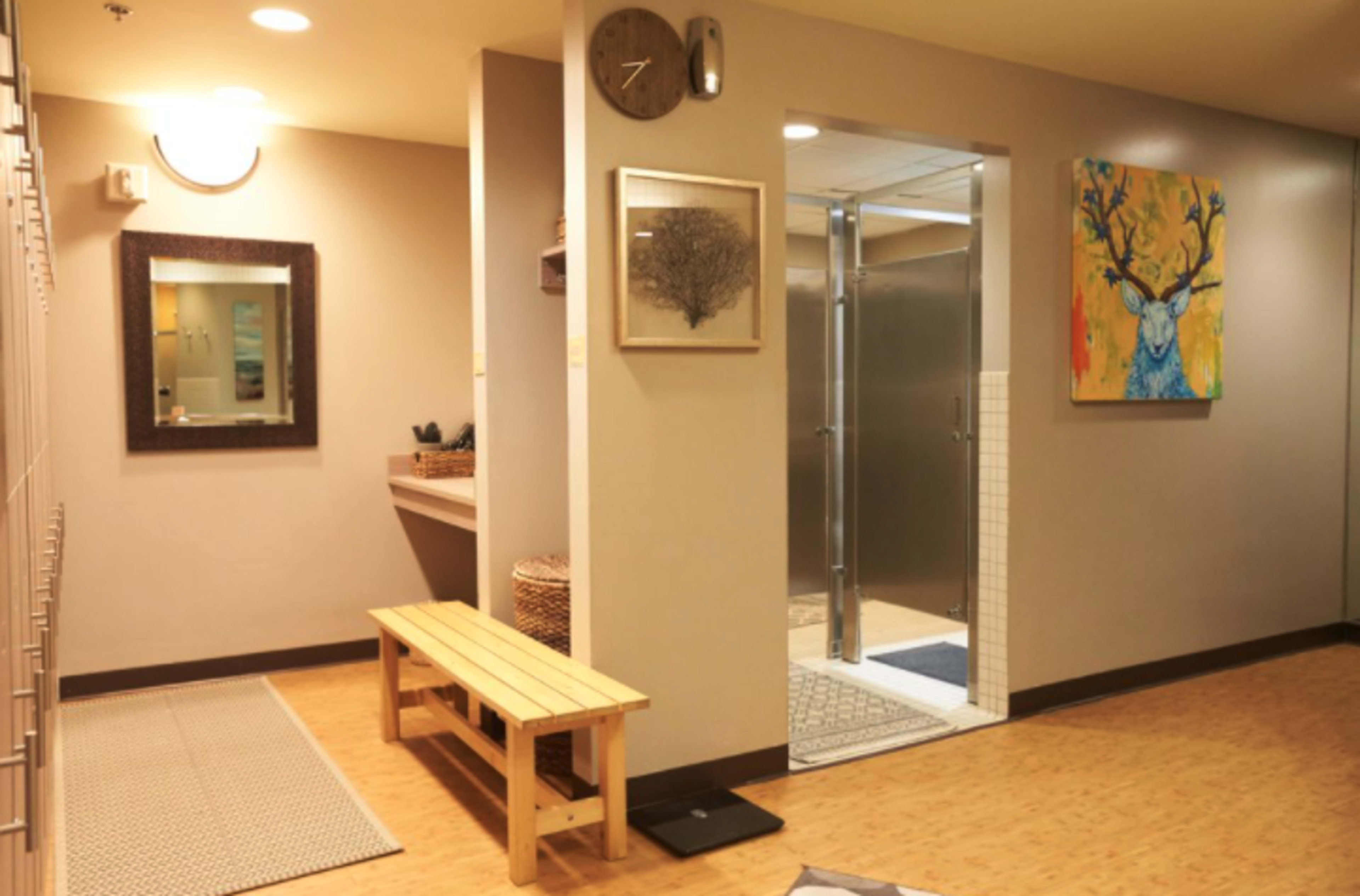 The image shows a locker room area with a wooden bench, wall-mounted artwork, and a glass-enclosed shower behind a tiled wall.