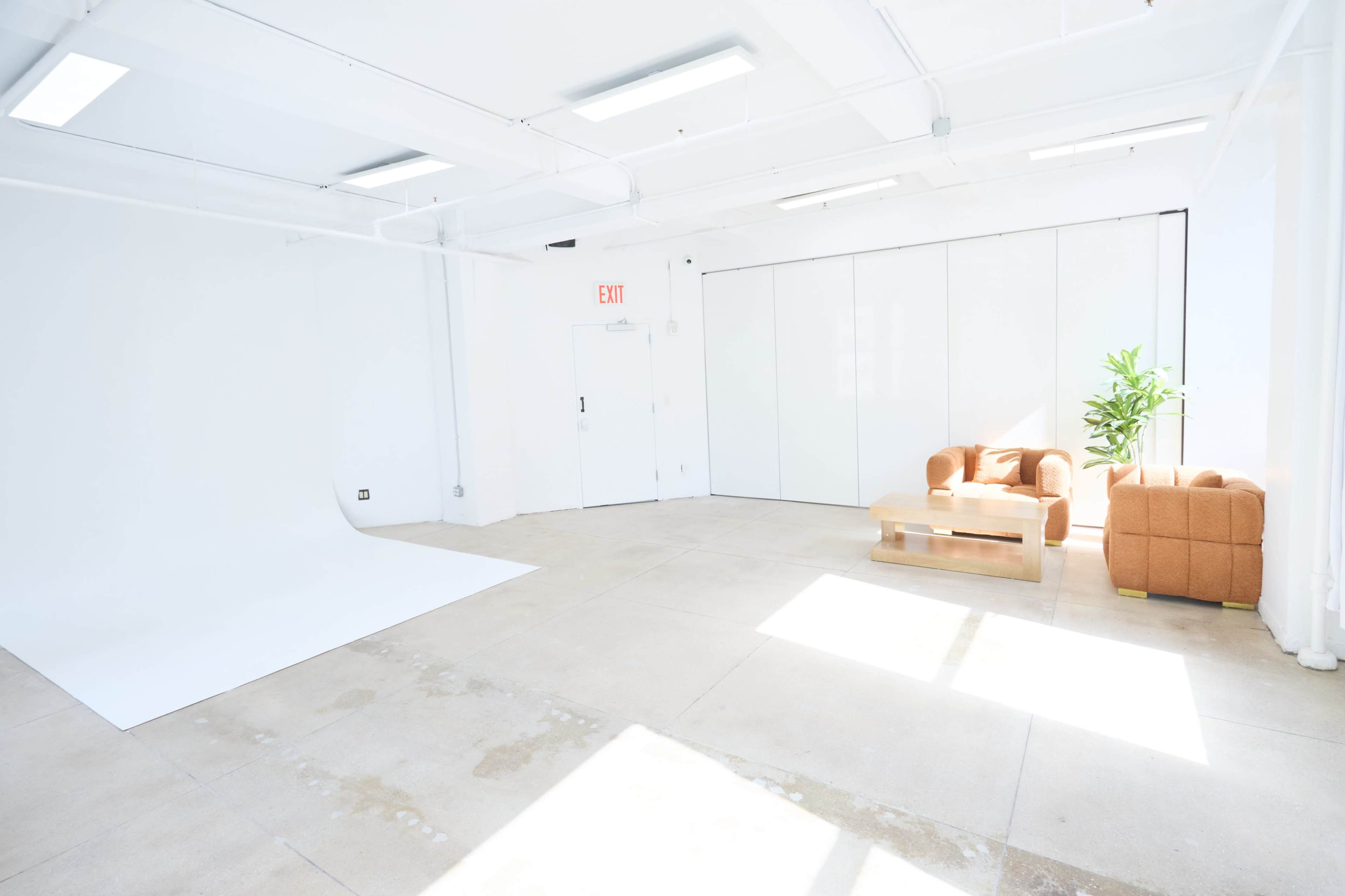 The image shows a bright, minimalistic studio space with a white backdrop, a wooden coffee table, and two tan chairs, along with a green plant in the corner.