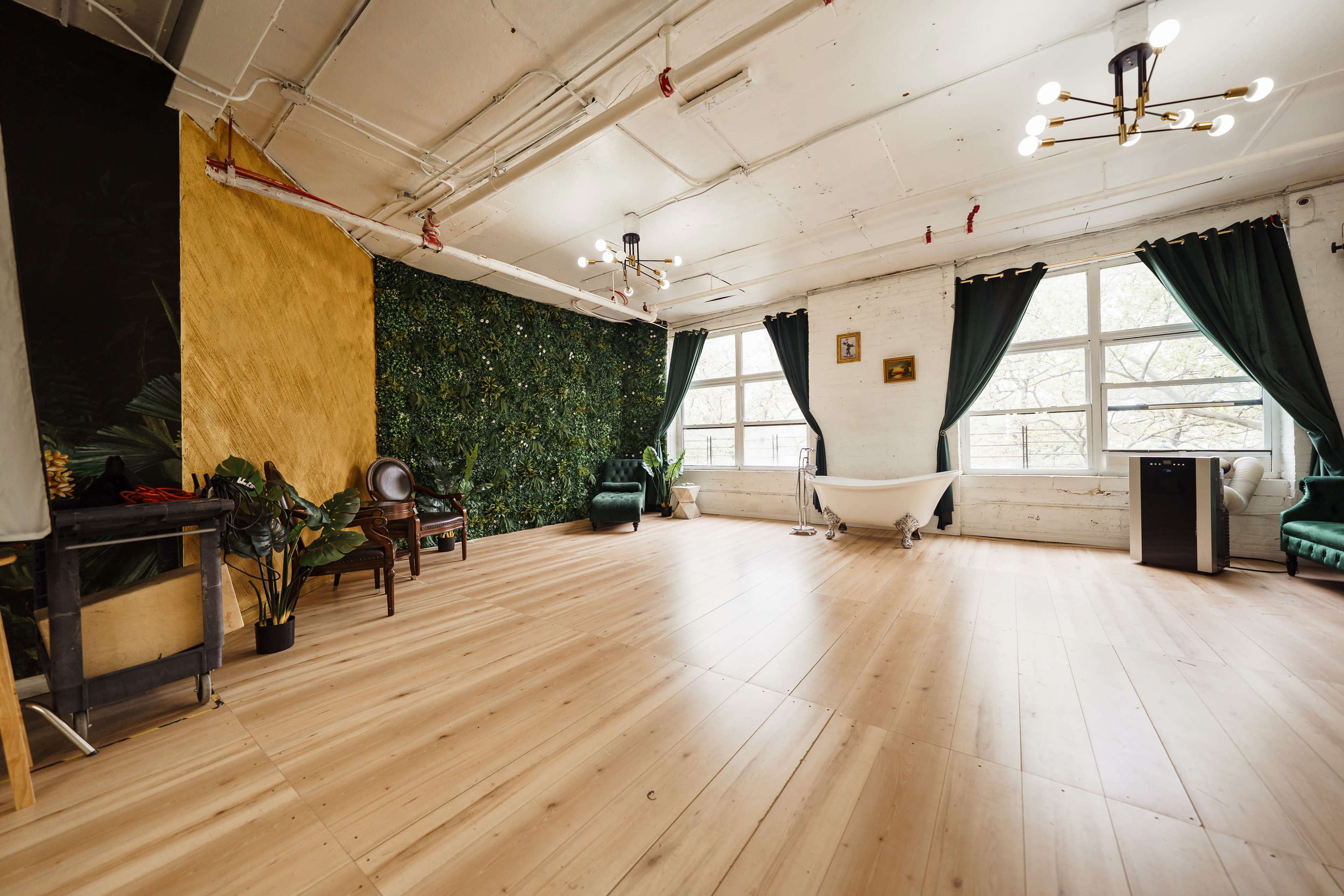 The image shows an expansive room with wooden flooring, featuring a green wall covered in plants, a gold-painted wall, a freestanding bathtub, and large windows with dark curtains.