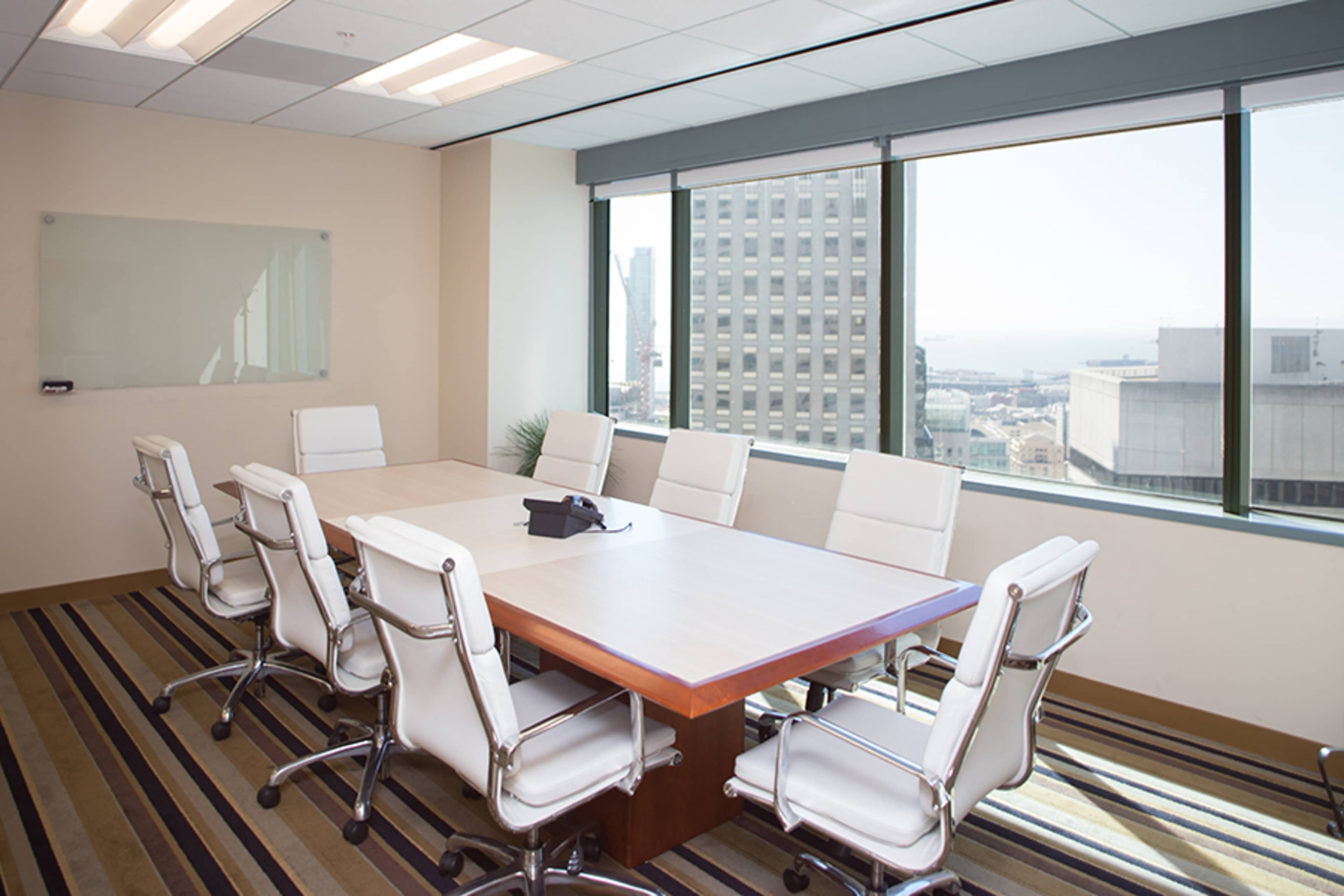 A modern conference room features a large wooden table surrounded by eight white ergonomic chairs and has windows providing a view of the city skyline.