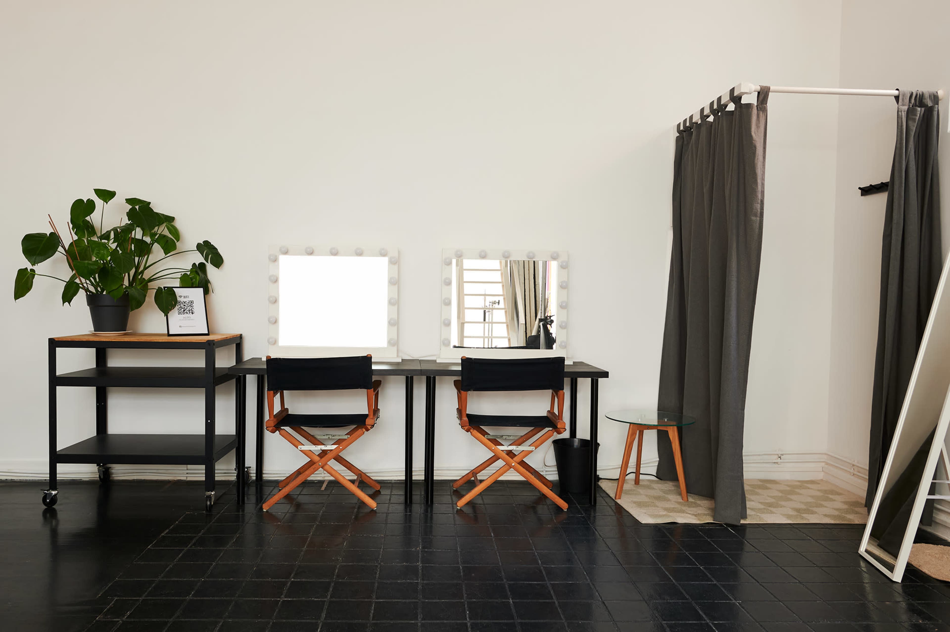 A minimalist makeup studio featuring two vanity mirrors with lights, two director's chairs, a potted plant, a cart, and a changing area with curtains.