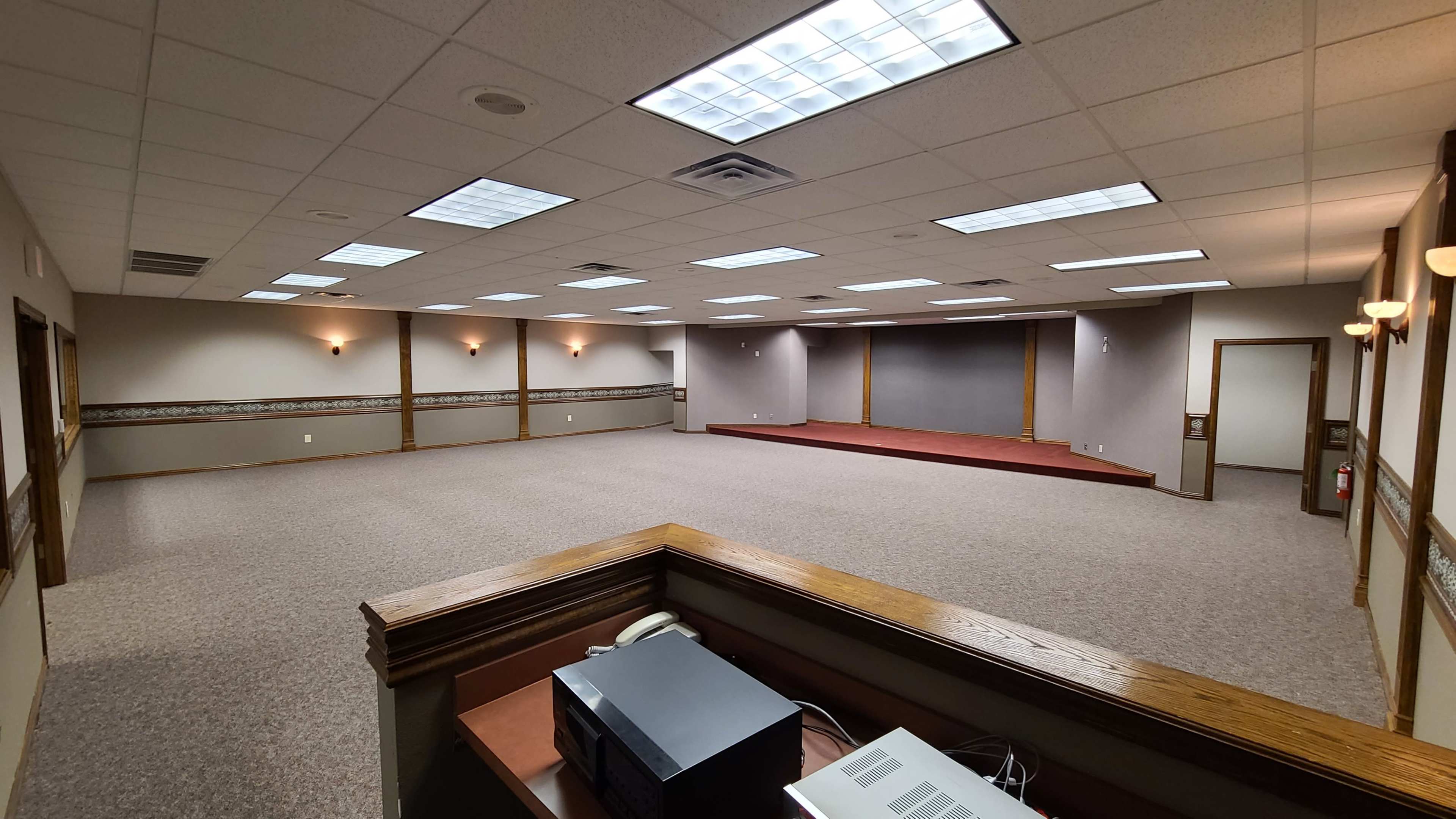A spacious, empty conference room with carpeted floors, overhead lighting, and a small stage at the far end.