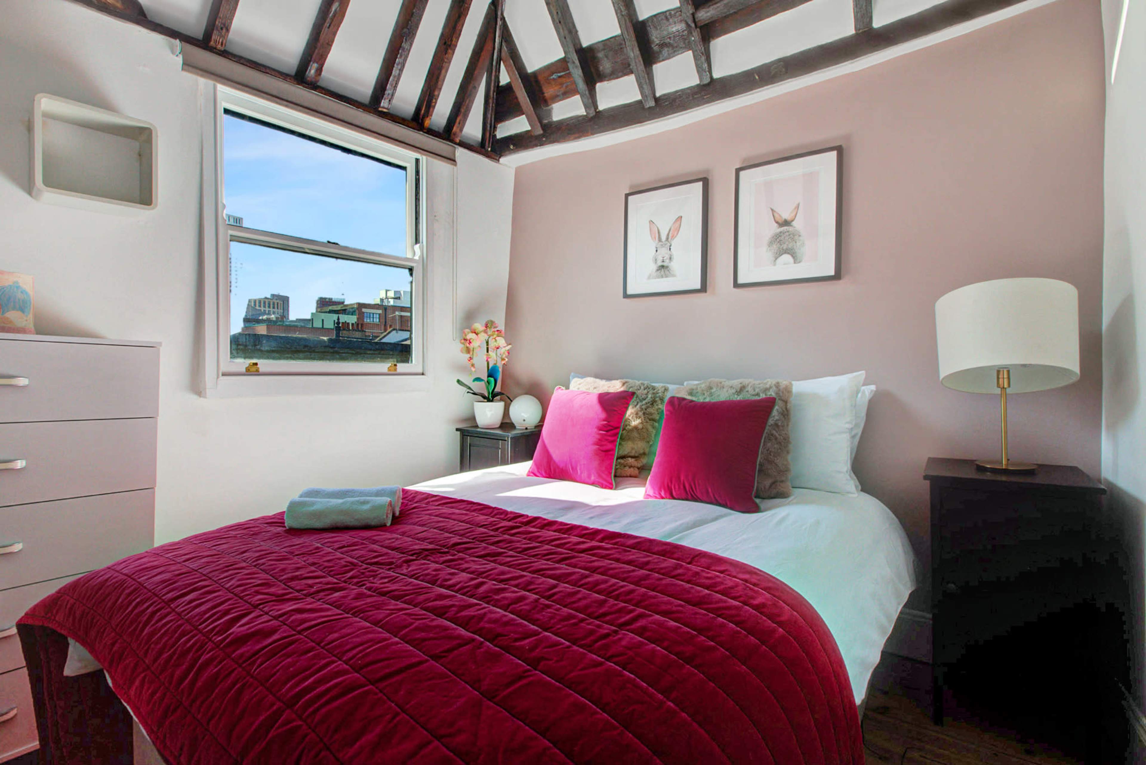 The image shows a bedroom with a bed covered in a burgundy quilt, two framed animal prints on the wall, and a window that lets in natural light.