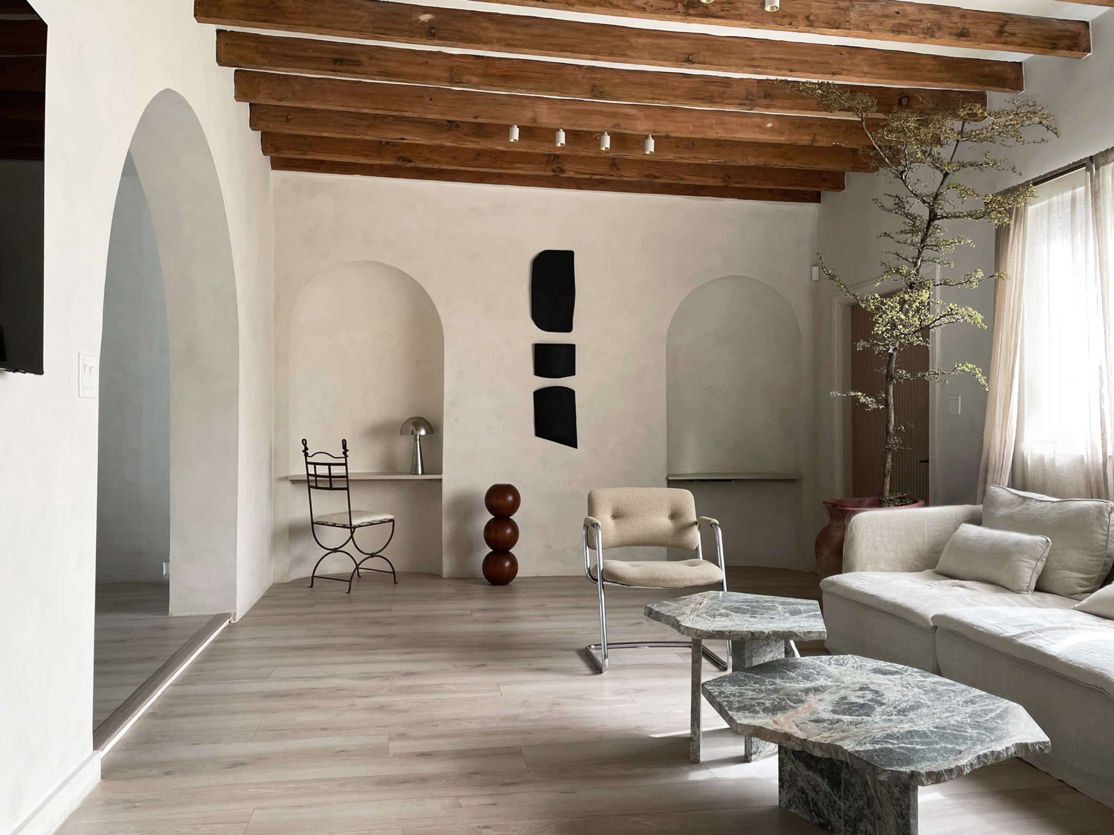 The room features a minimalist design with wooden beams, a beige sofa, a round stone table, and a vertical arrangement of artworks on the wall.