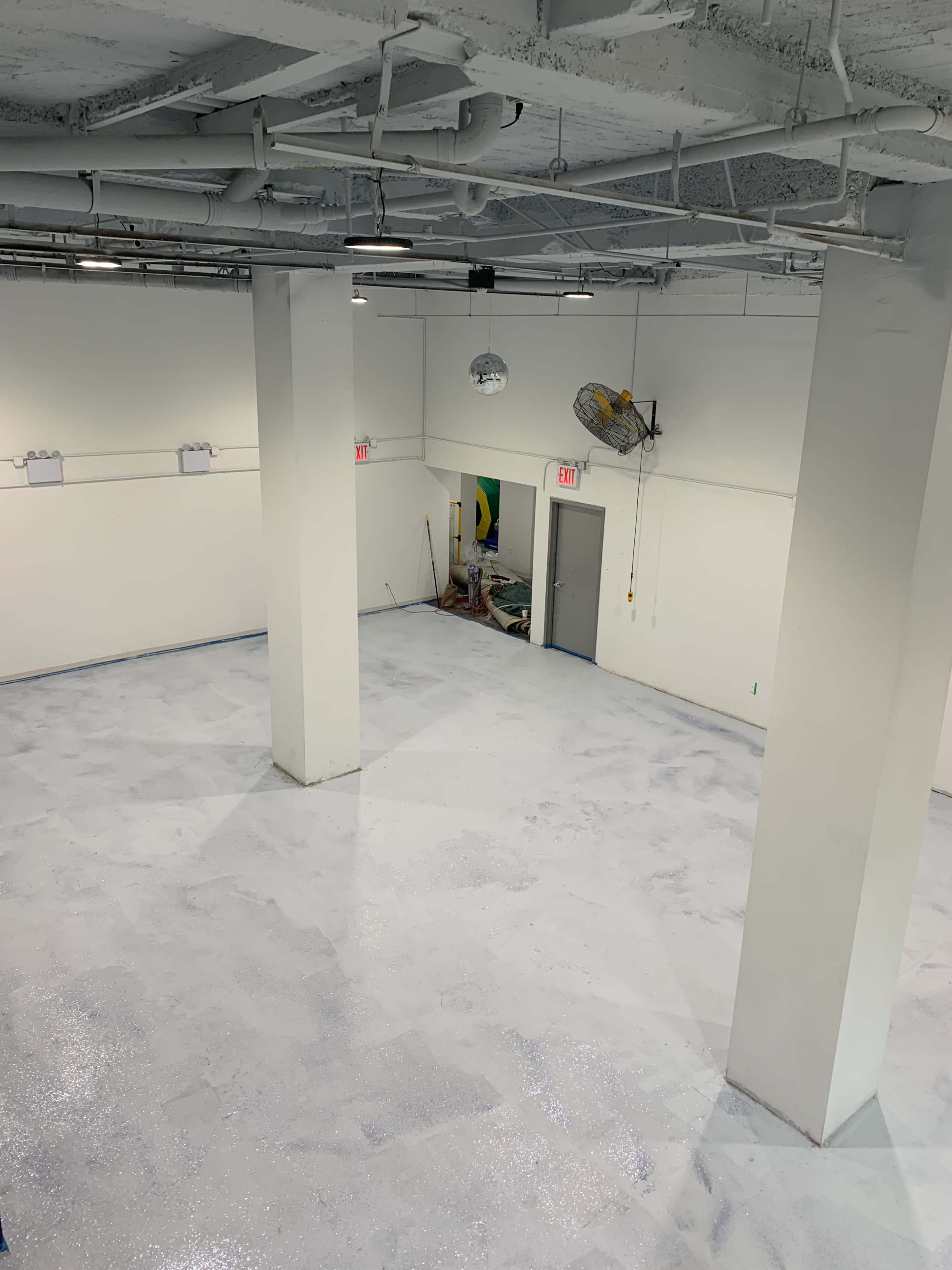 The image shows a large, empty room with a polished, shiny floor and white walls, featuring several ceiling-mounted lights and an open doorway at the far end.