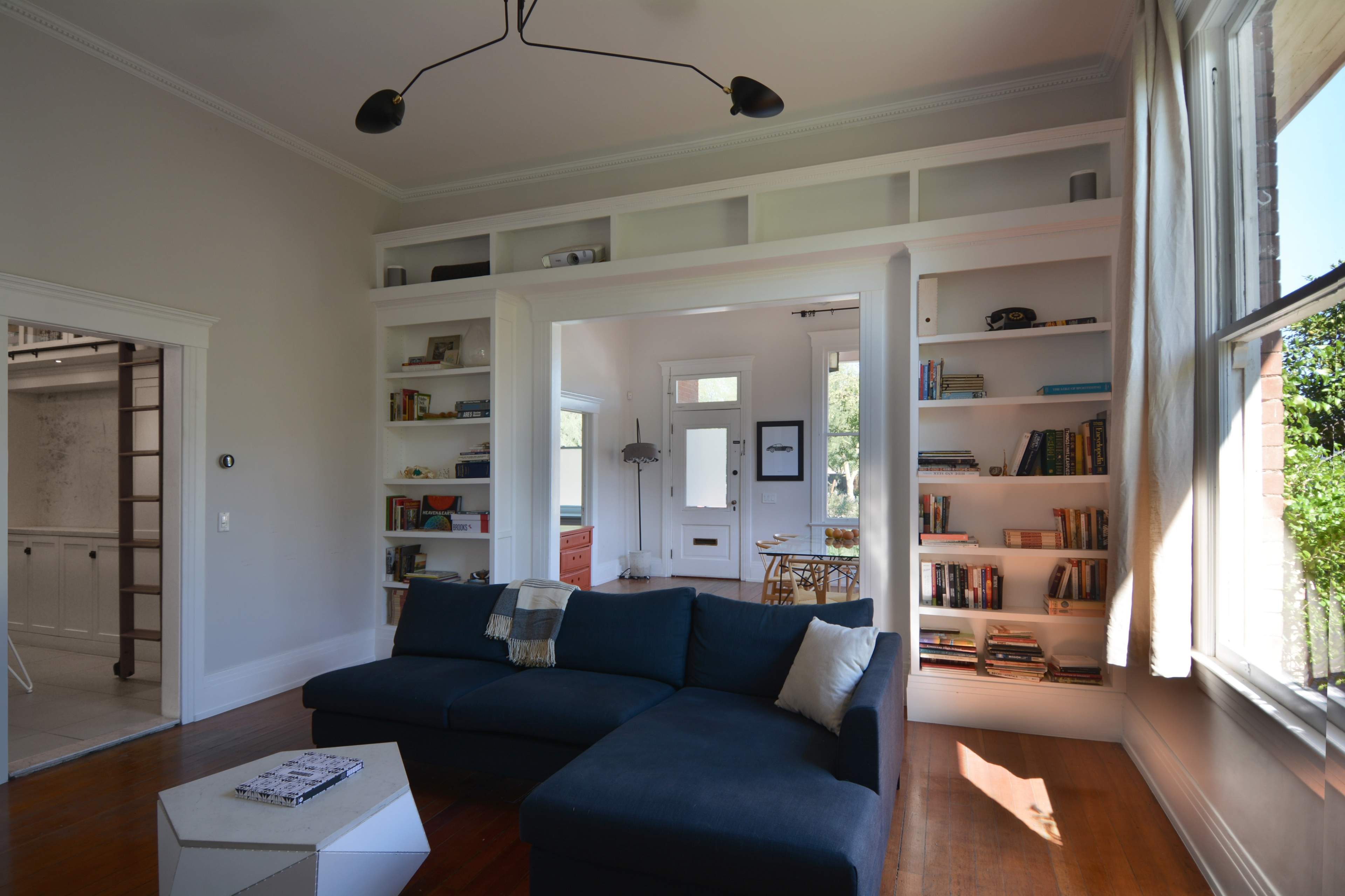 A bright living room with a dark sectional sofa, built-in shelves filled with books, and large windows allowing natural light to enter.