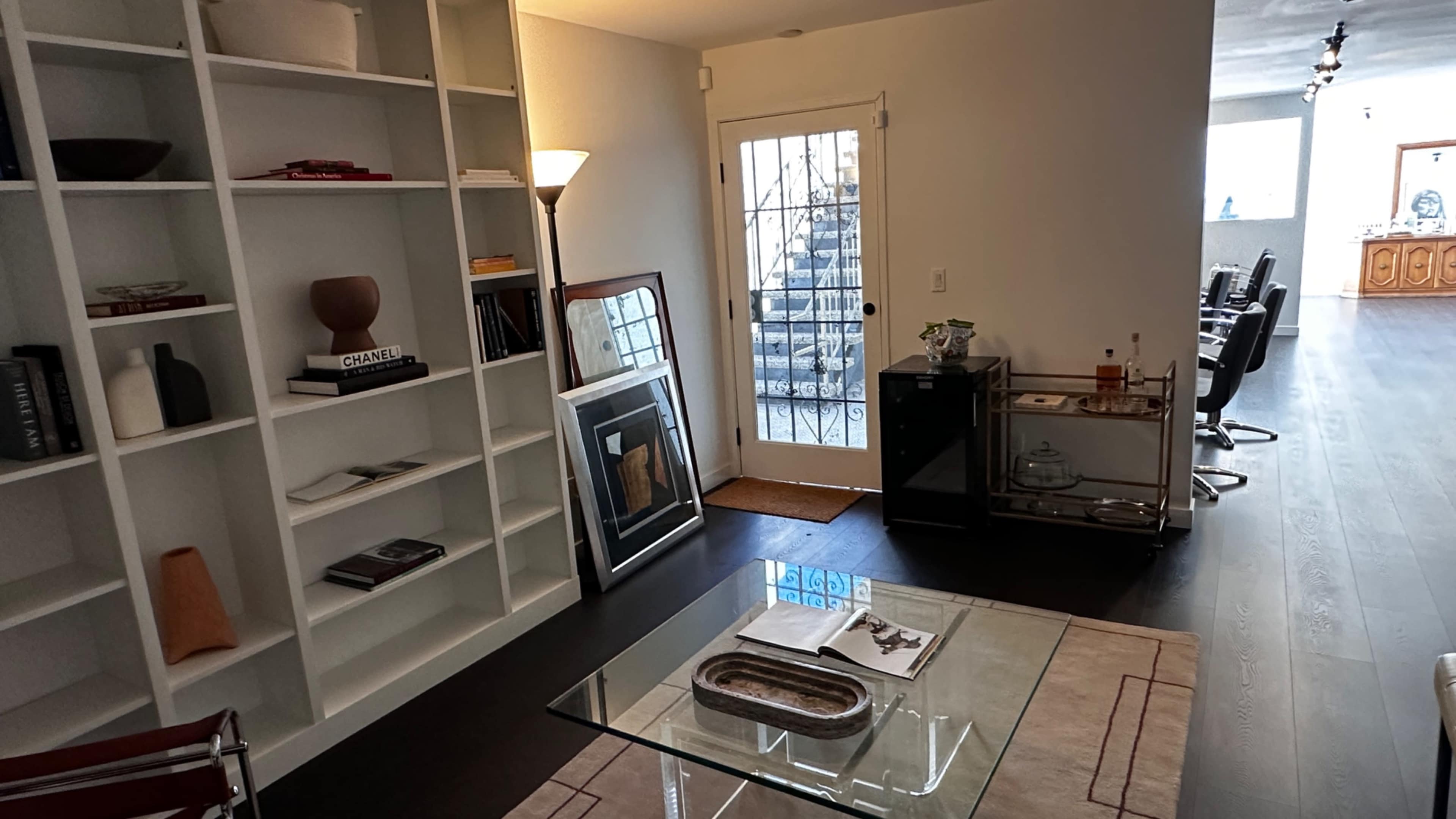 A modern living area with a glass coffee table, shelving unit filled with decor items, and a door leading to an outside area.