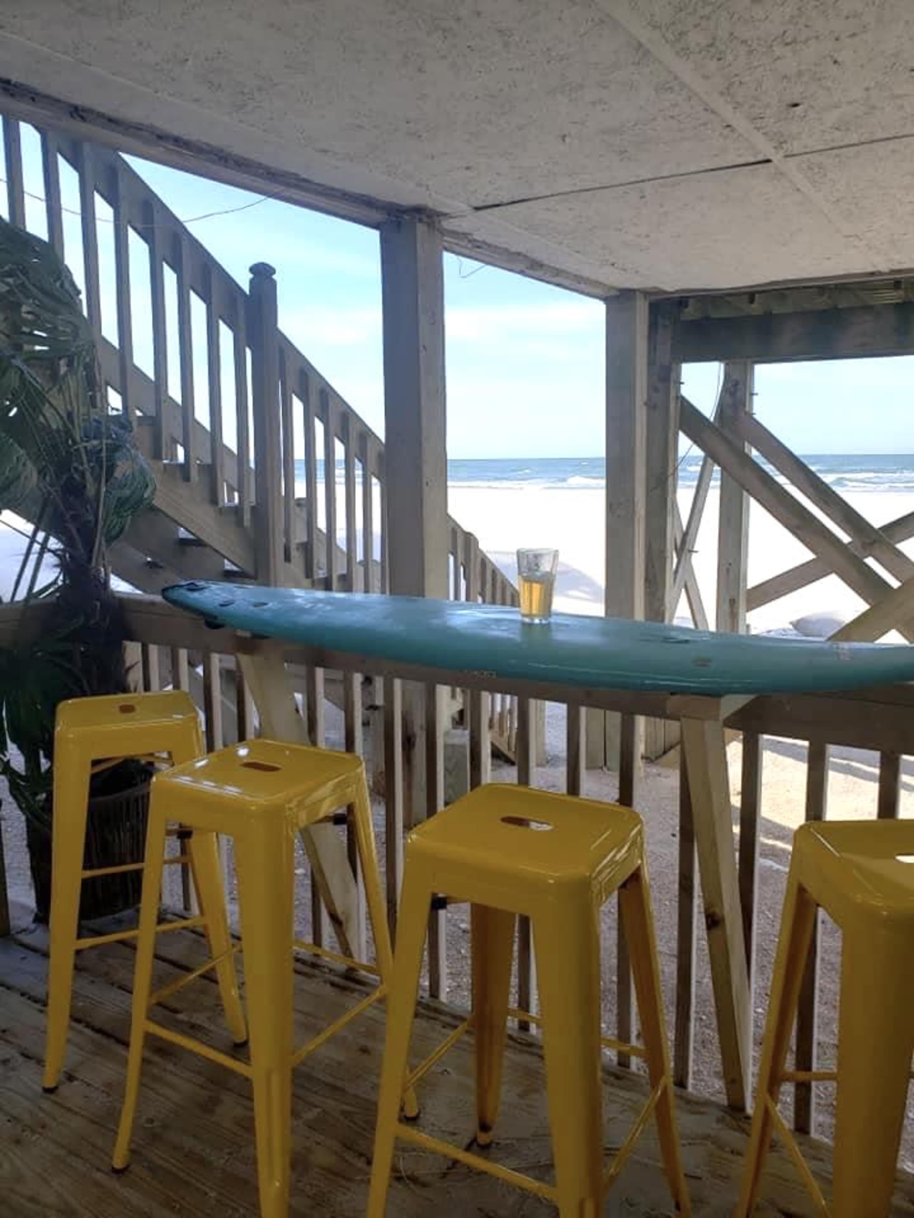 A row of yellow bar stools sits in front of a turquoise bar overlooking a sandy beach and ocean.