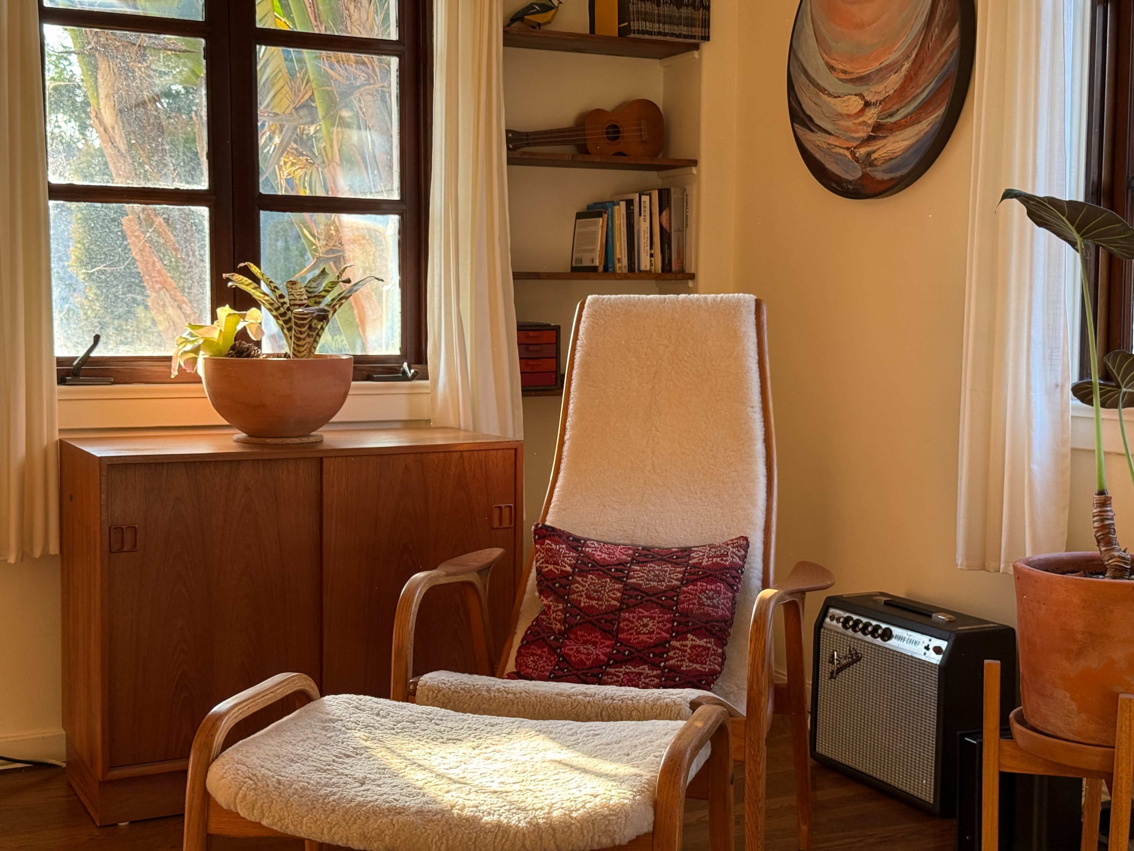 A cozy sitting area features a wooden chair with a patterned cushion, a small footrest, a potted plant, and a bookcase in a sunlit room.