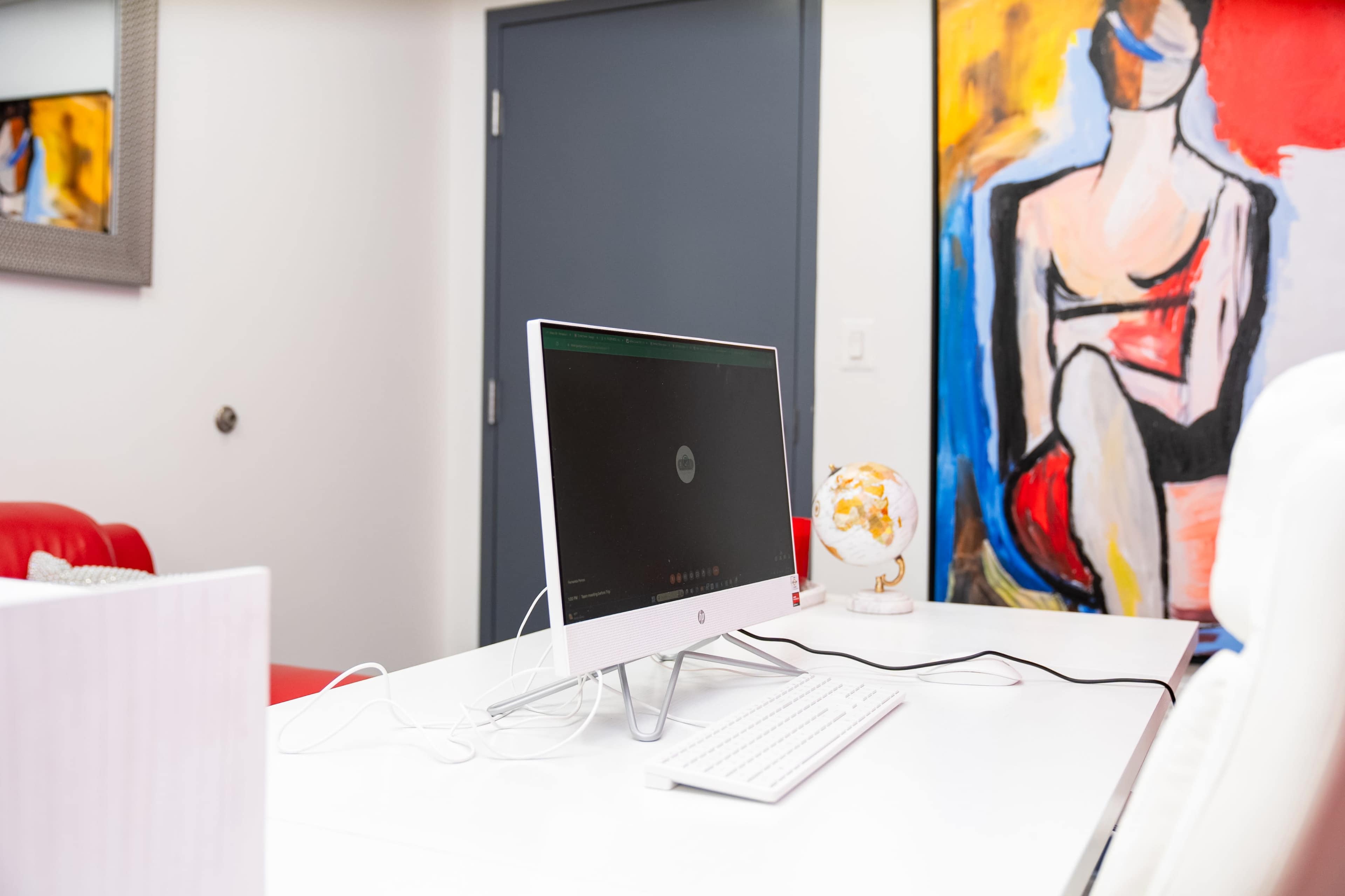 A sleek desktop computer is positioned on a white desk next to an artistic painting and a decorative globe.