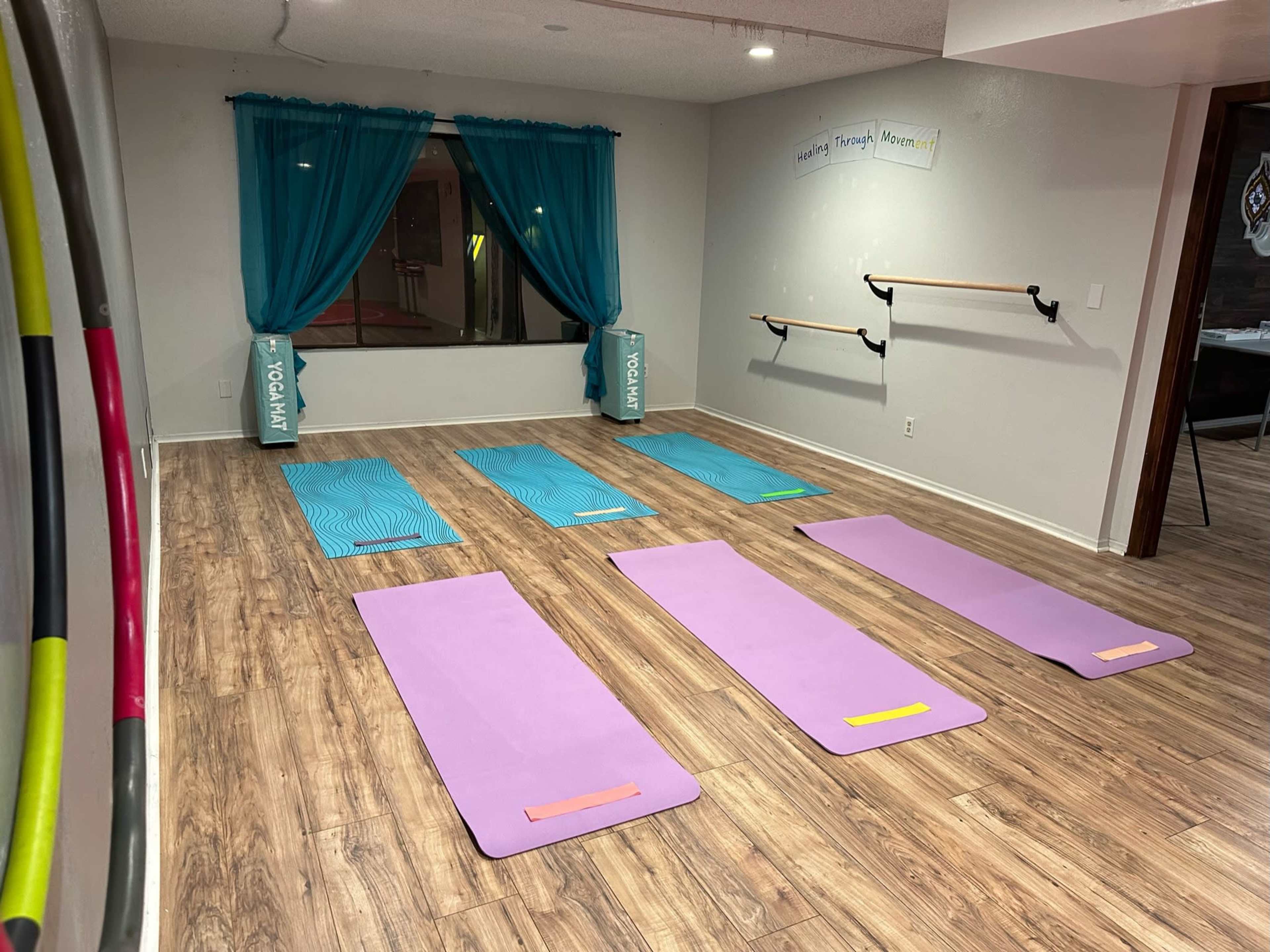 A clean exercise studio with six yoga mats arranged on a wooden floor, along with blue curtains and barre equipment on the walls.
