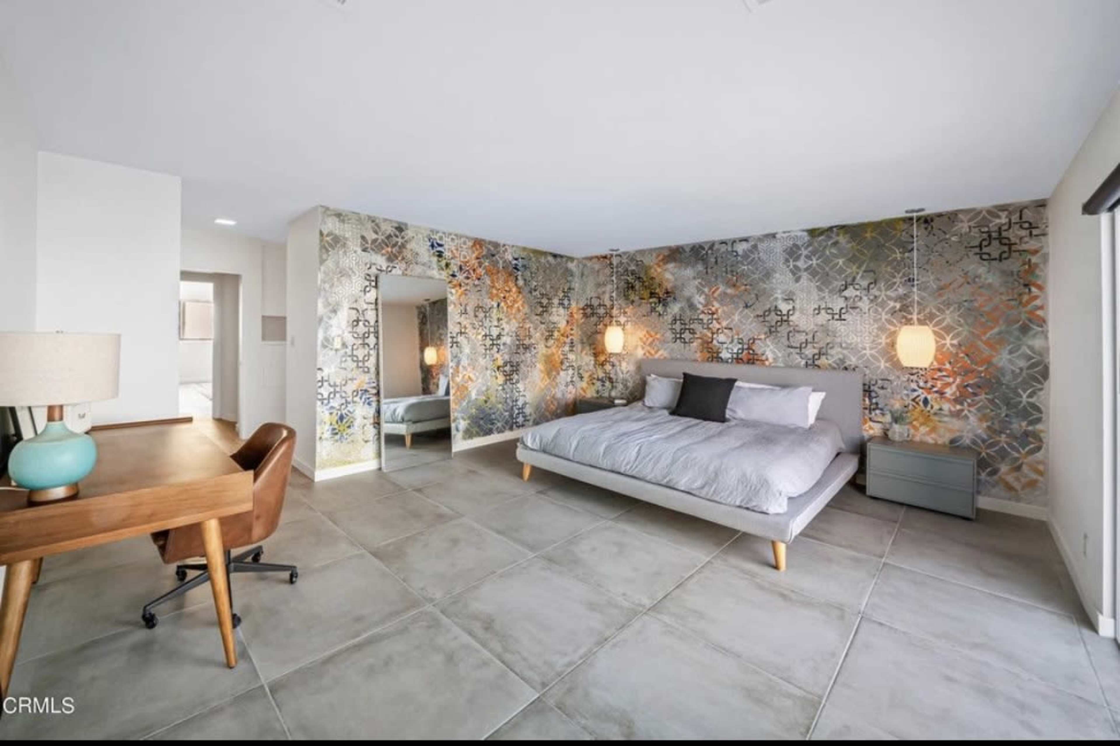 A spacious bedroom features a large bed, a distinct patterned accent wall, and a mirror beside a wooden desk and chair.