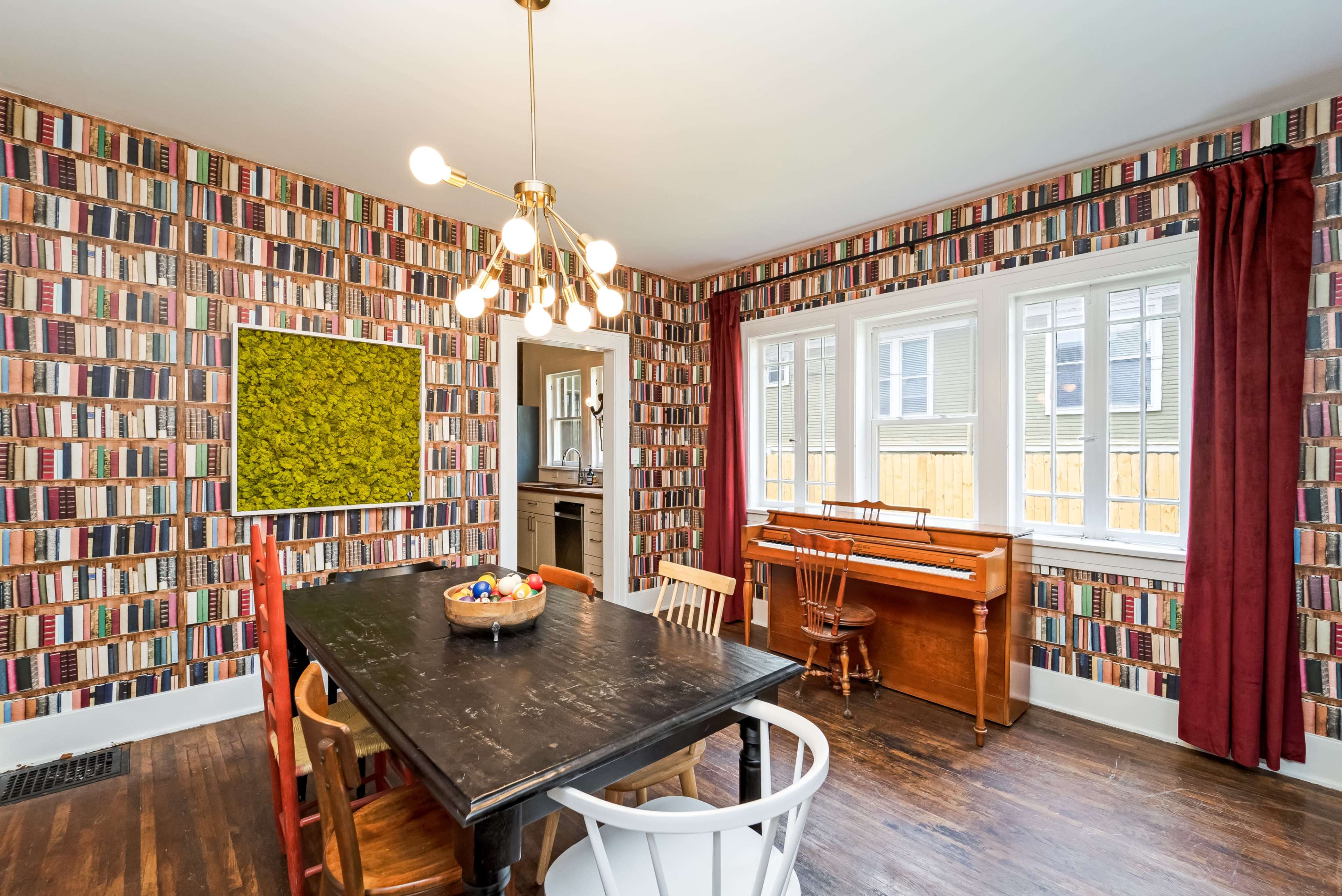 The dining area features a table surrounded by chairs, a piano against the wall, and wallpaper designed to resemble bookshelves.