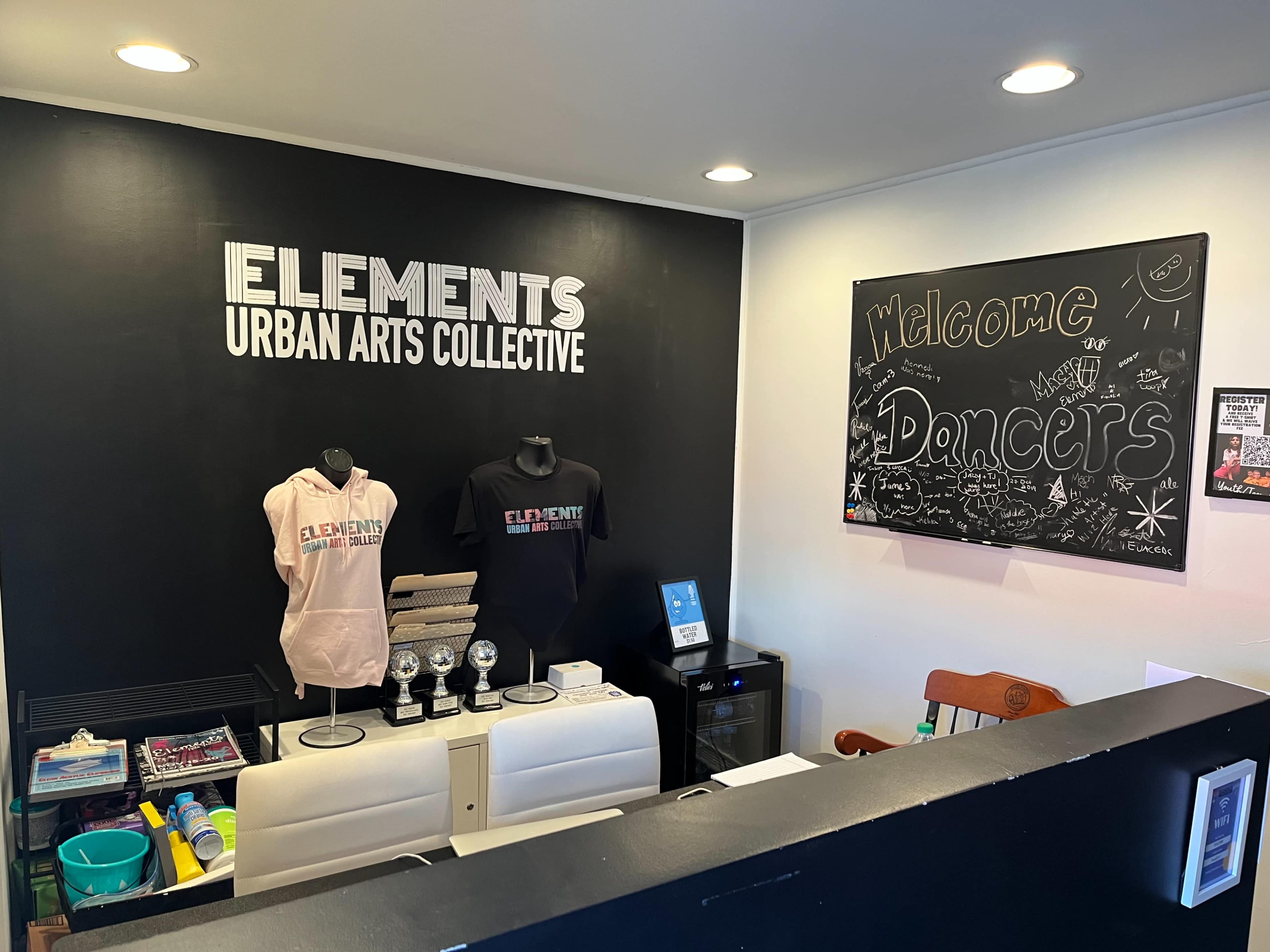 A reception area with merchandise display for the "Elements Urban Arts Collective," including branded apparel and a welcome message for dancers on a chalkboard.