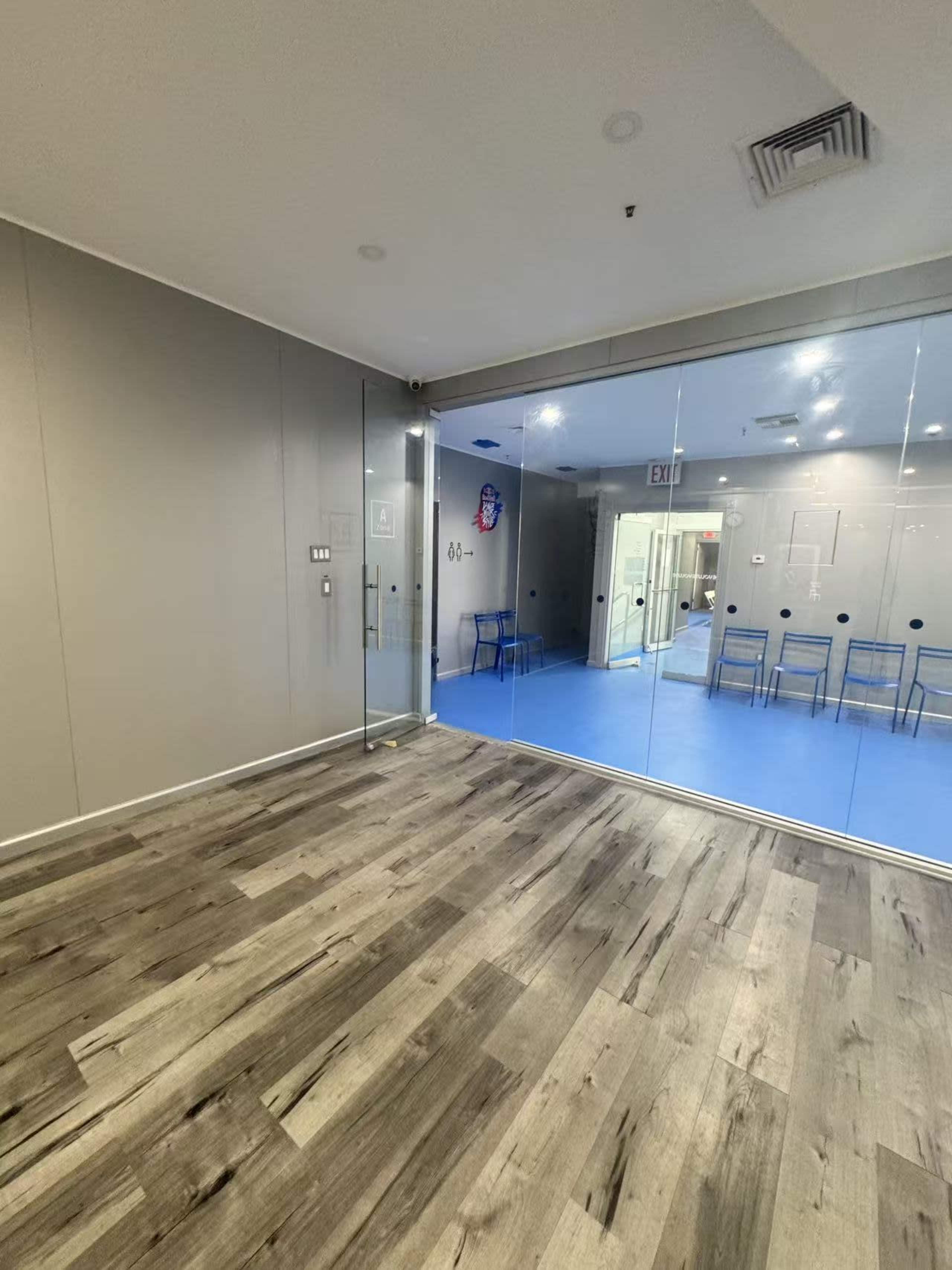 The image shows a modern interior space with a wood floor and a large glass wall separating two areas, one of which features blue flooring and several chairs.