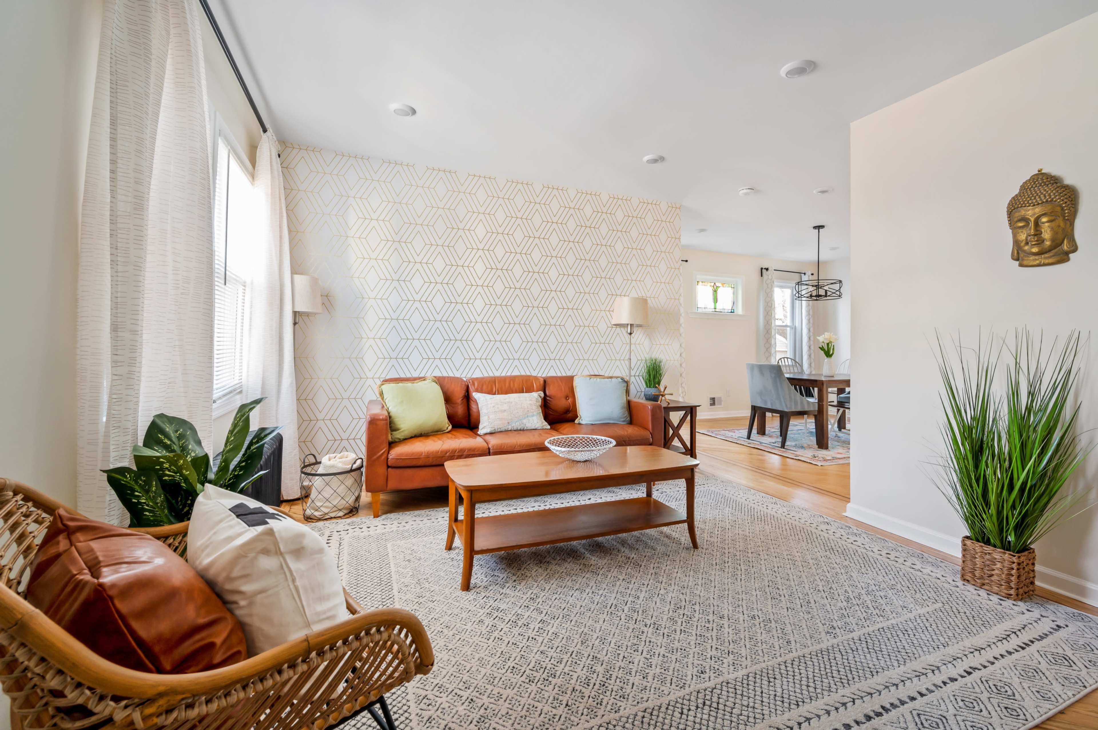 A cozy living room featuring a brown sofa with colorful cushions, a wooden coffee table, and decorative plants, complemented by patterned wallpaper and a woven chair.