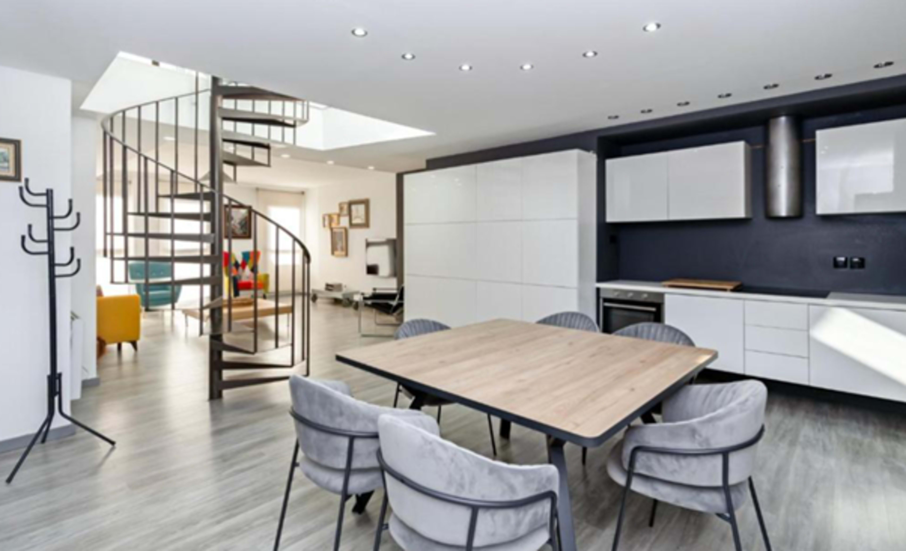 A modern open-concept interior features a dining area with a large wooden table surrounded by upholstered chairs, leading to a spiral staircase and a sleek kitchen.