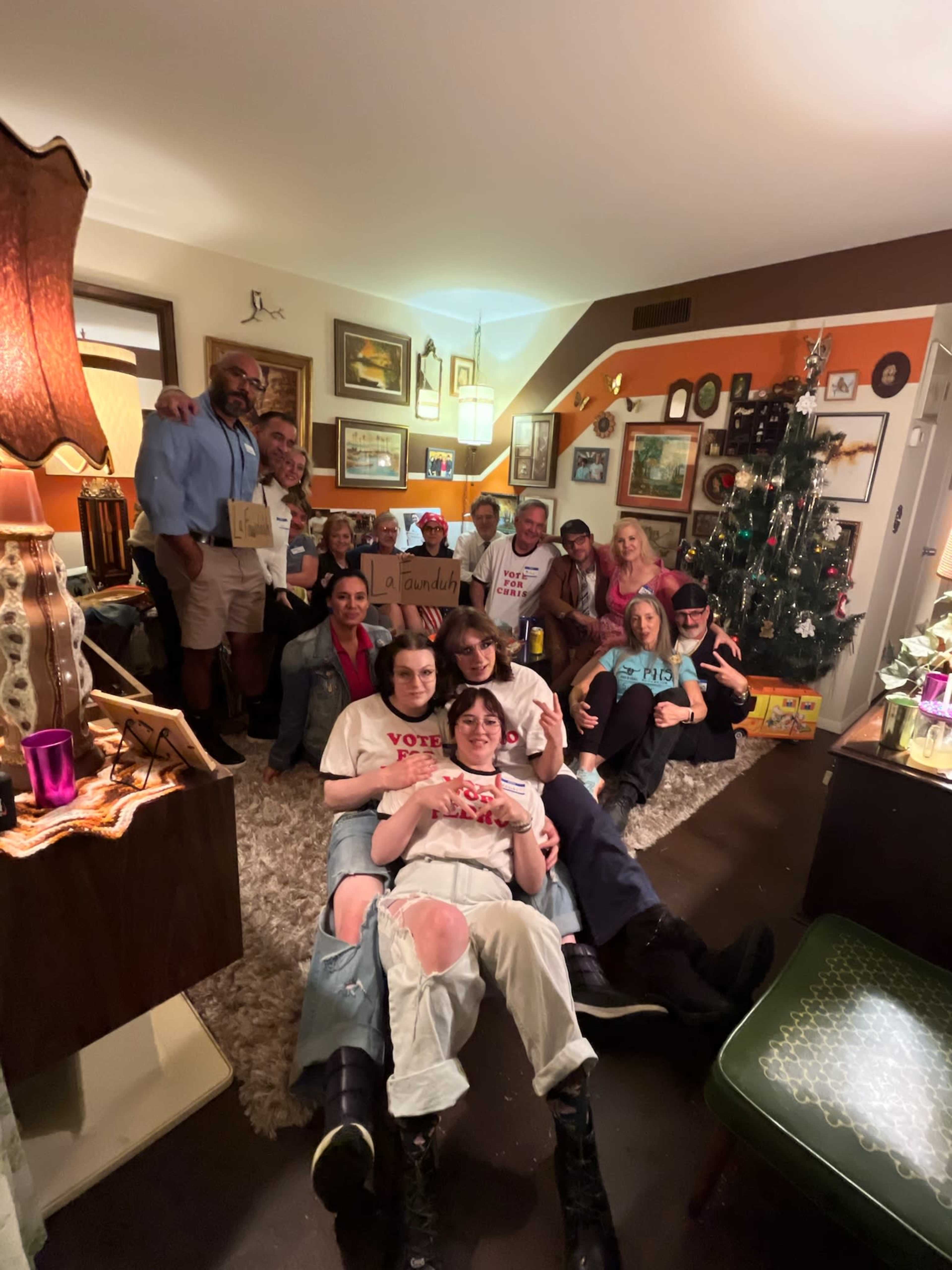 A diverse group of people is gathered in a cozy living room, posing for a photo with festive decorations and a Christmas tree in the background.