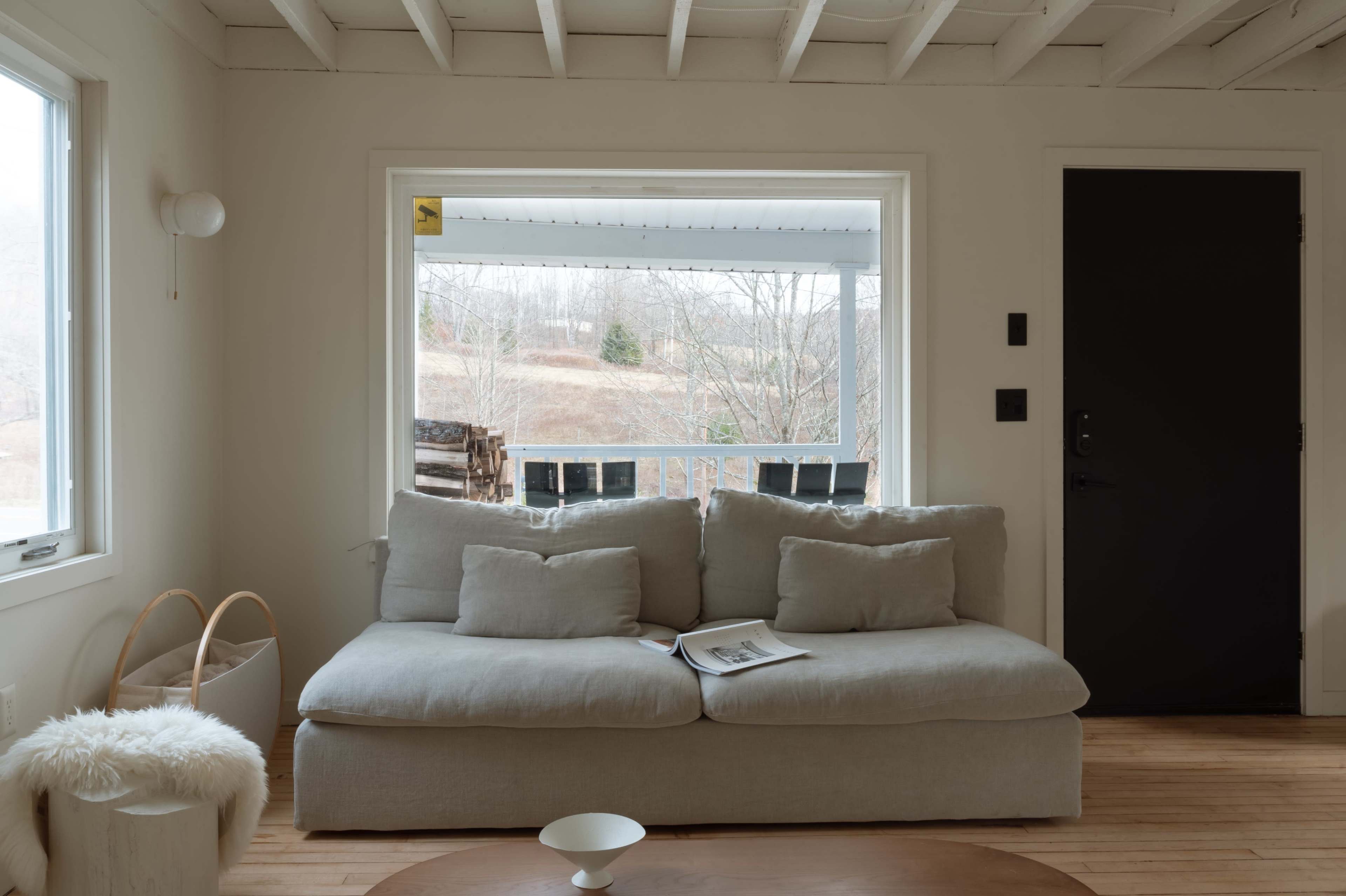 A light-colored living room with a large, comfortable sofa facing a window that overlooks a hillside landscape outside.