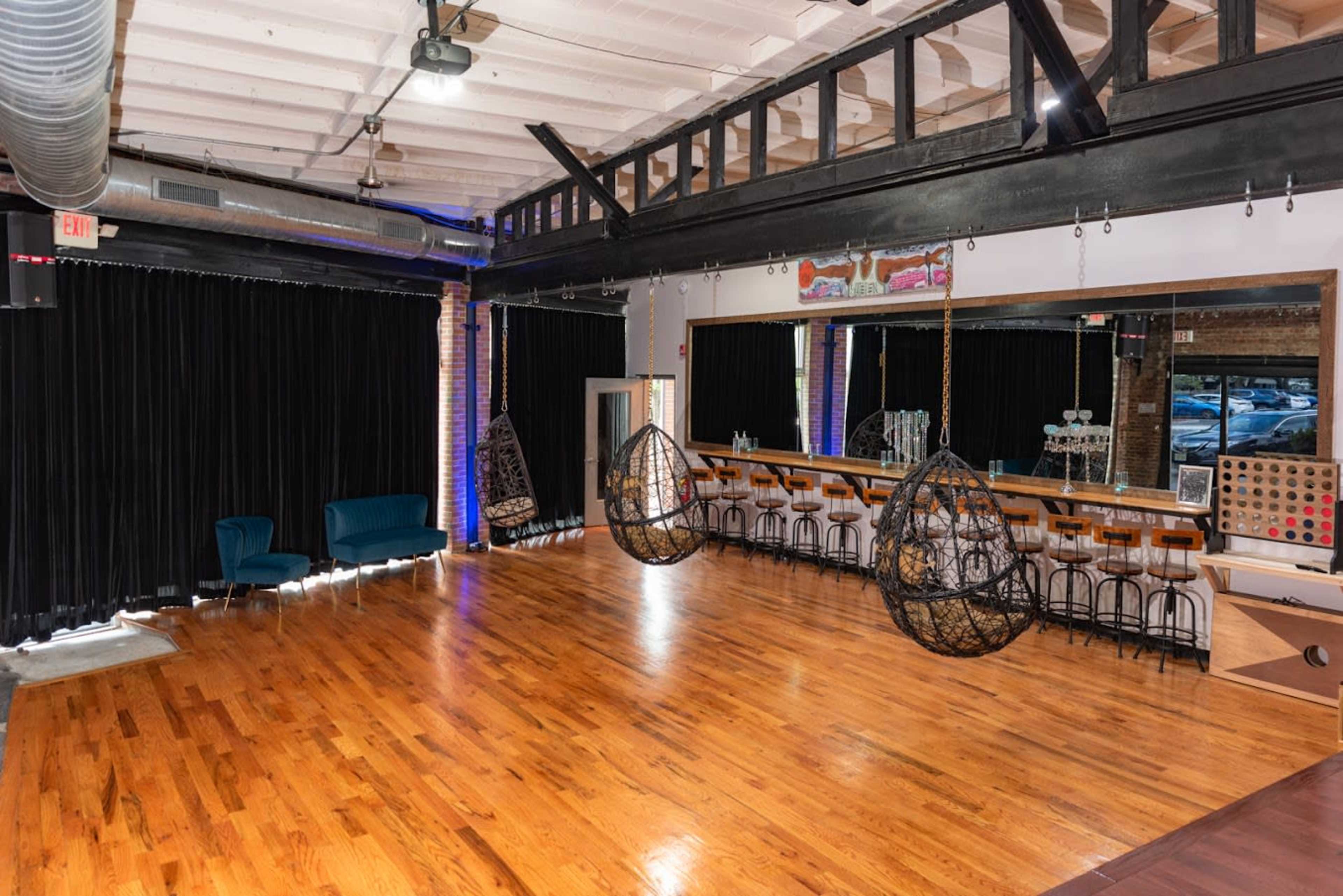 The interior space features wooden floors, hanging chairs, a bar with stools, and black curtains along the walls.