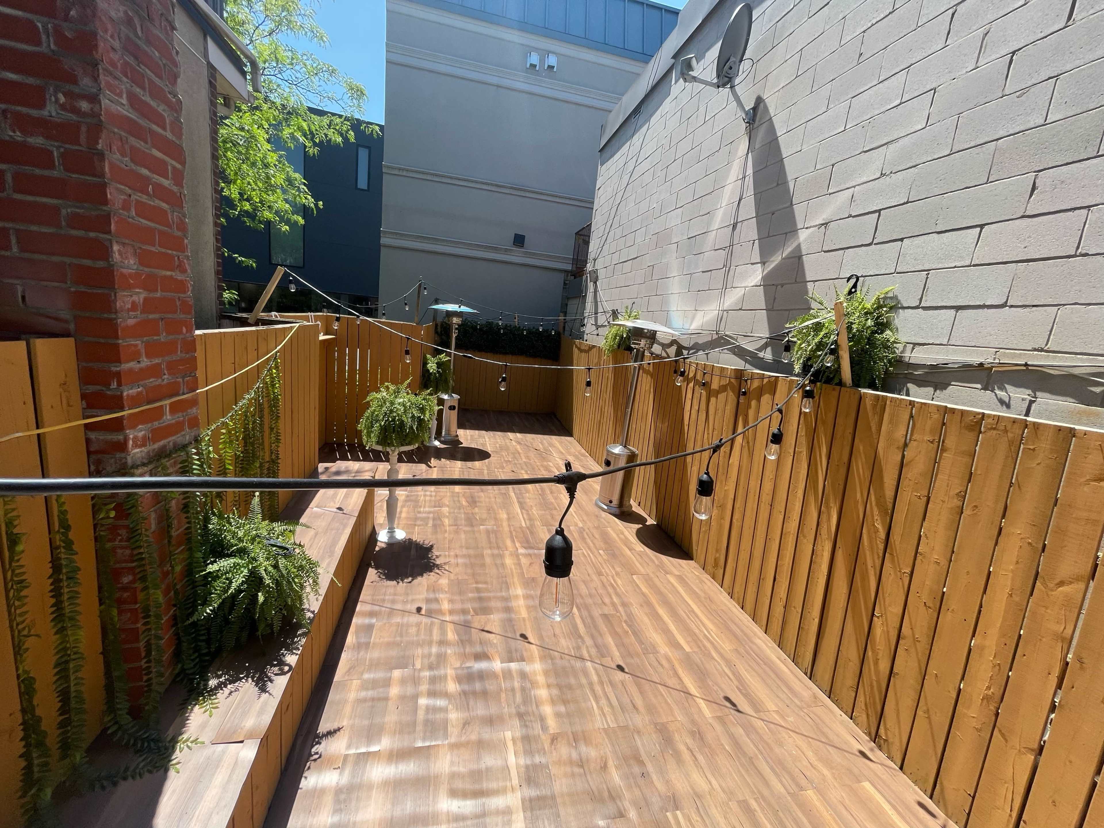 The image shows a sunlit wooden patio area with hanging lights and greenery along wooden fencing.