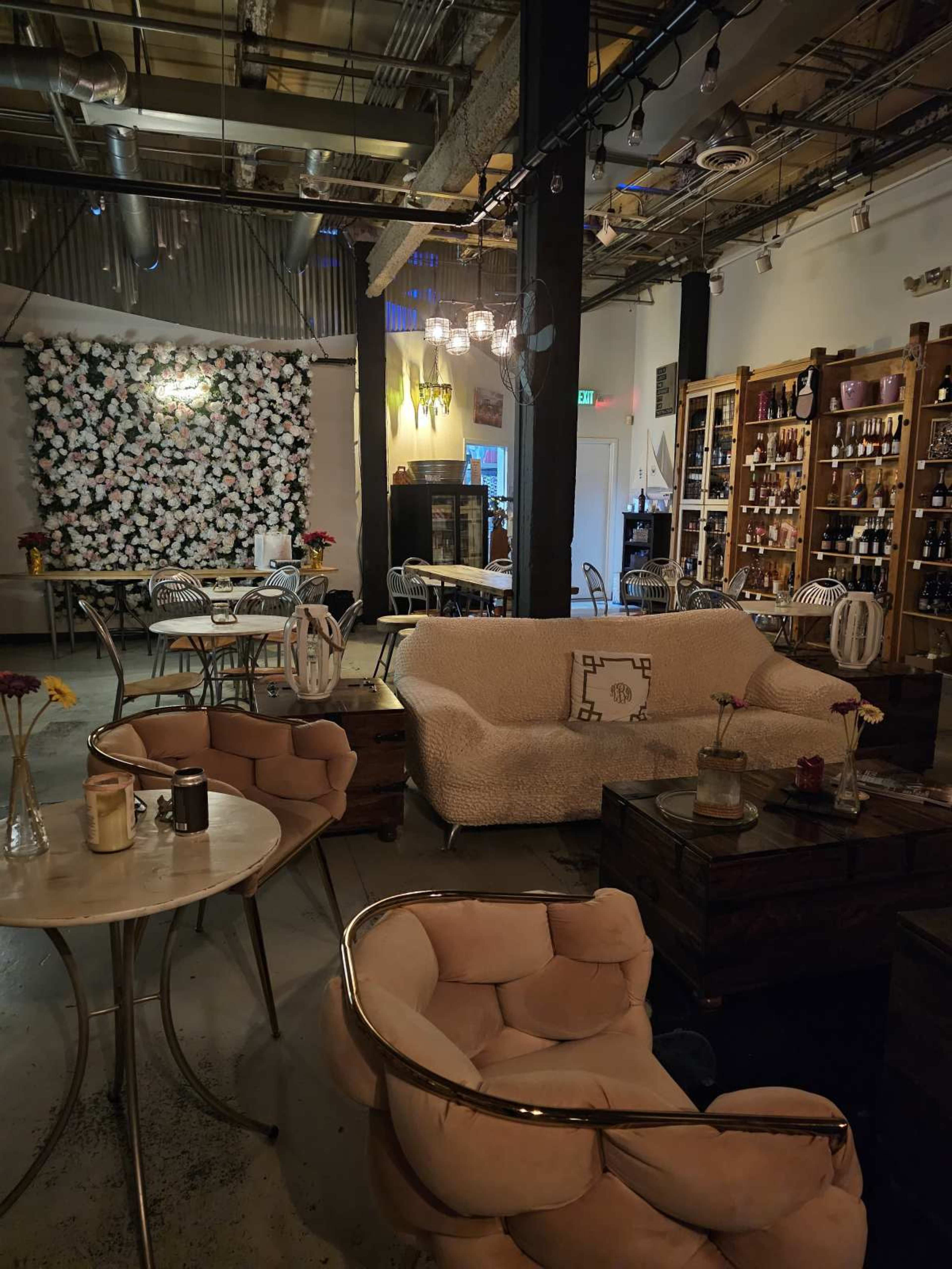 The image shows a cozy café interior with floral wall decor, seating arrangements including a white sofa and two gold-accented chairs, and shelves filled with bottles.