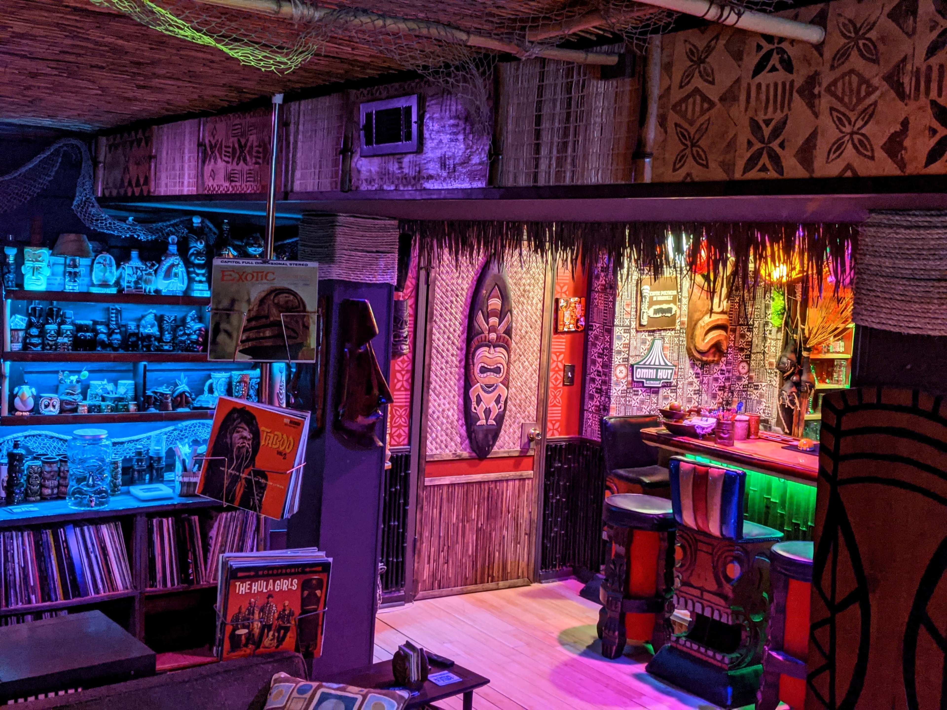 The image shows a dimly lit tiki bar with colorful lighting, featuring a wooden counter, tiki masks on the walls, and shelves filled with tropical-themed decor and vintage records.