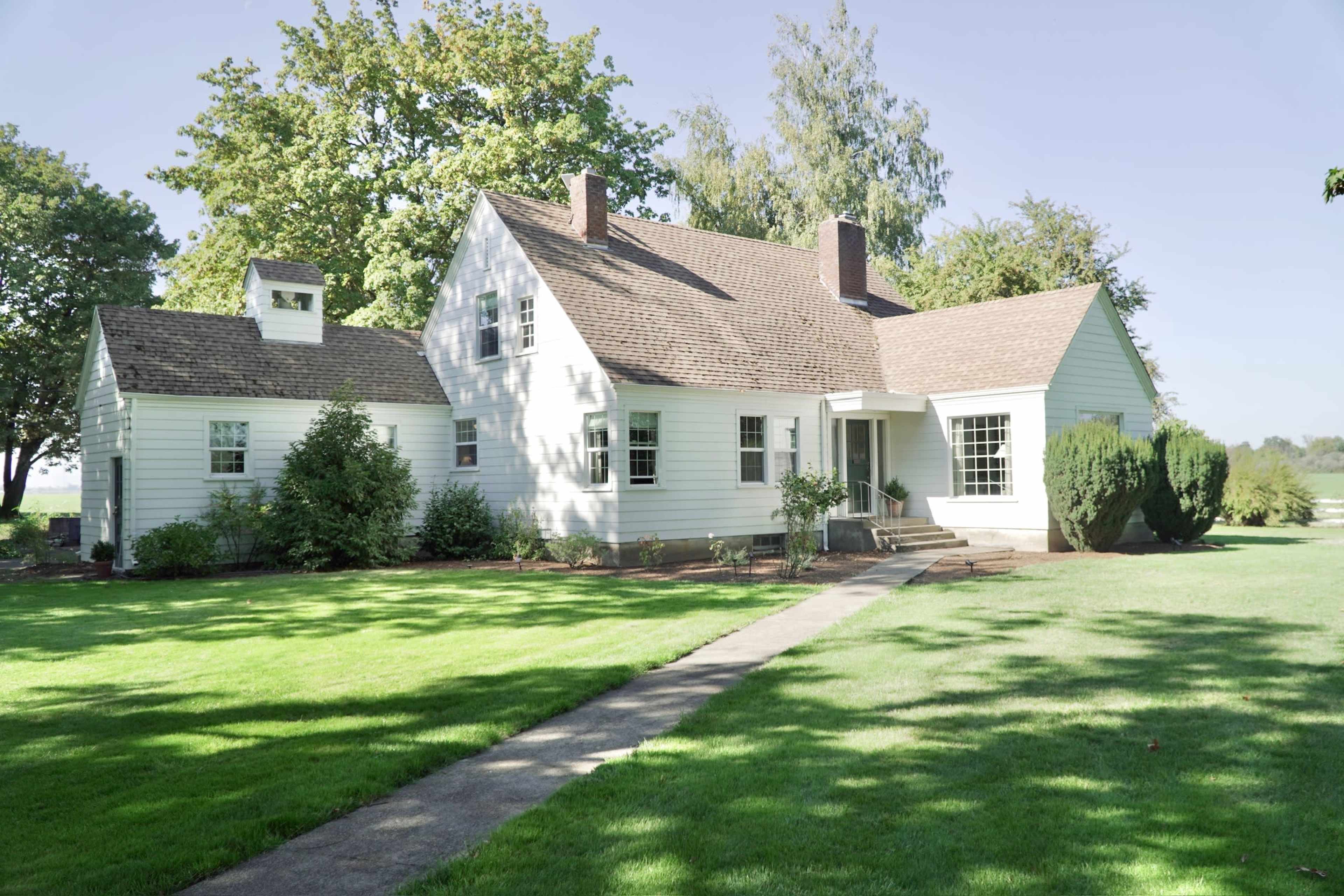 A white, two-story house with a sloped roof, surrounded by green grass and trees, with a concrete pathway leading to the front entrance.