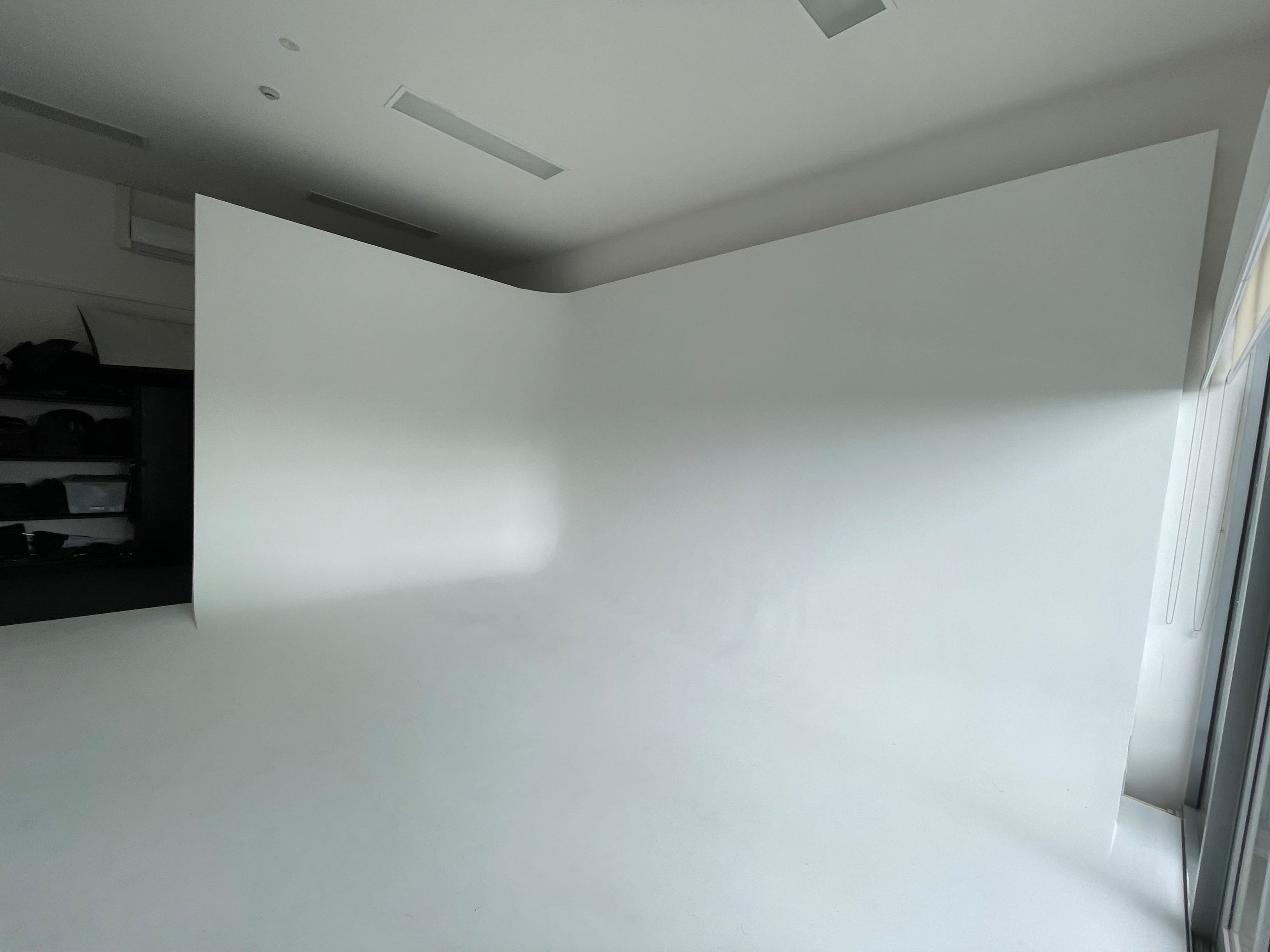 A spacious, empty room with a seamless white backdrop and a partially visible dark storage area in the corner.