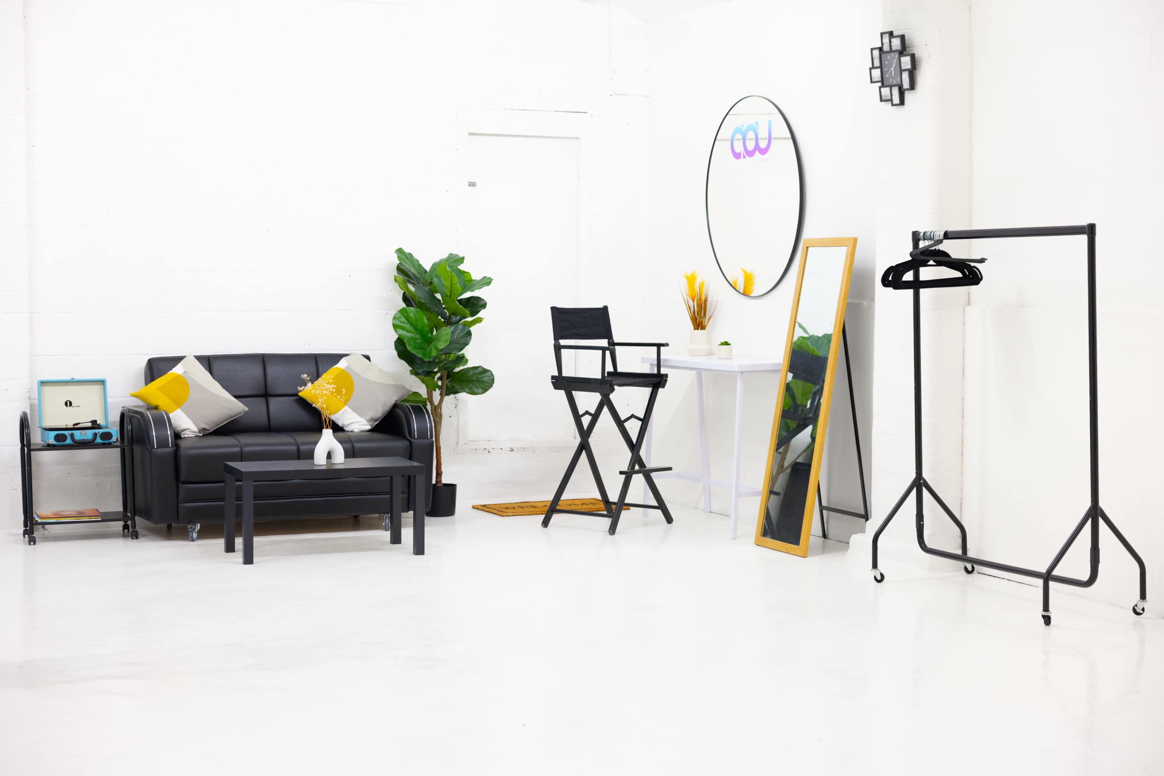 The scene features a modern, minimalist studio space with a black couch, a director's chair, a tall mirror, a clothing rack, and some decorative plants.
