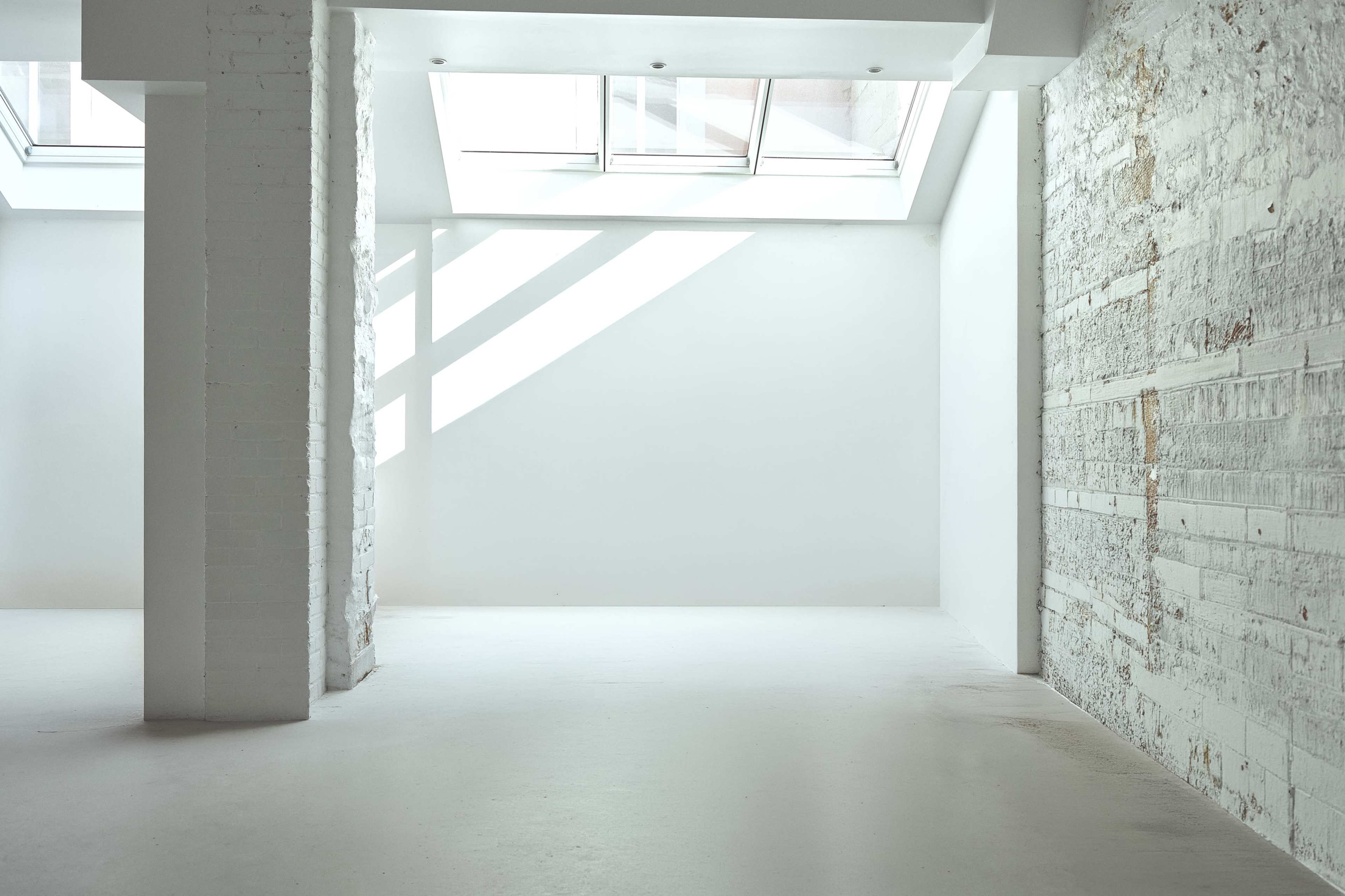 The image shows a spacious, minimalist room with white walls, large windows allowing natural light, and a polished floor.